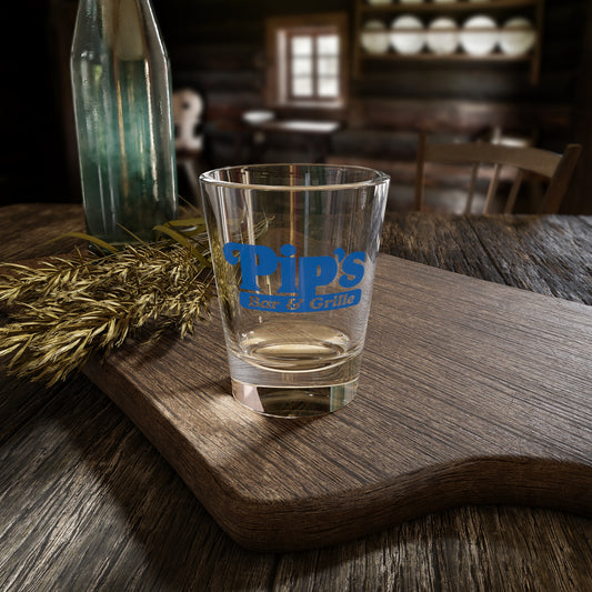 Mockup of a 1.5 oz ounce clear shot glass featuring the Pips Bar and Grill logo from the Apple TV show Severance printed on one side, shown on a wooden table in a kitchen.