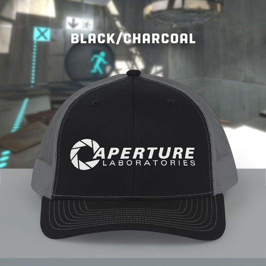 Front view of a black/charcoal colored Richardson 112 Snapback Trucker Cap featuring the embroidered logo of the fictional company Aperture Laboratories from the video game Portal and Portal 2, sold by ScriptedBrand.