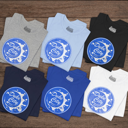 Six folded t-shirts with vintage distressed Duke City Dairy logo inspired by TV Show Pluribus, displayed flat wood background.