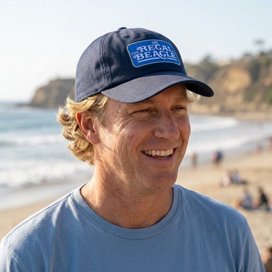 Man at a beach wearing Navy blue dad cap with embroidered Regal Beagle patch logo, Three's Company inspired fictional bar merch