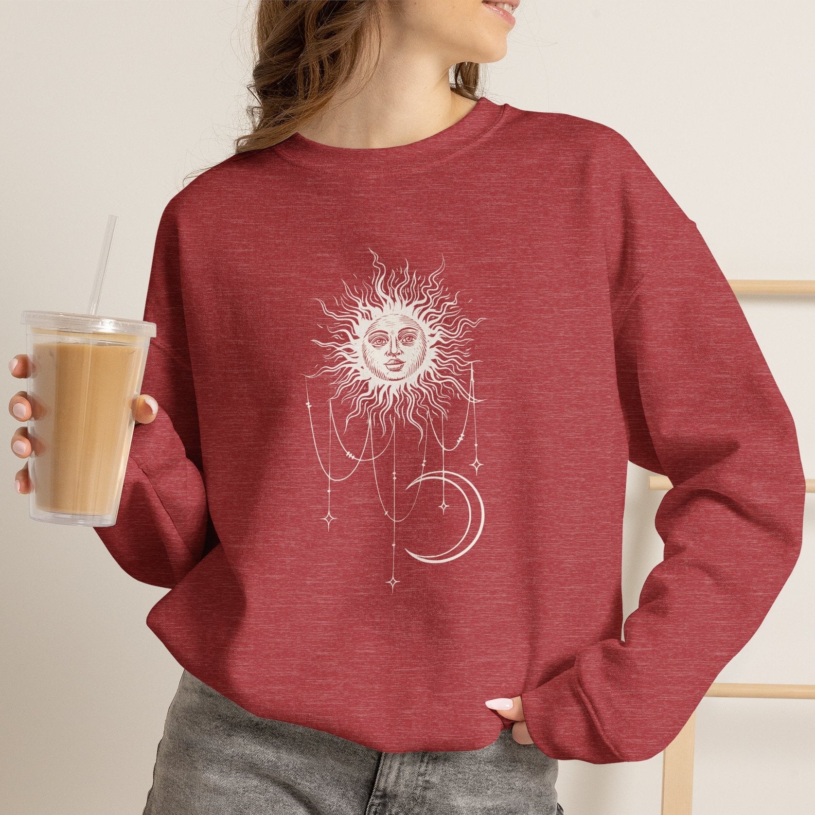 Person wearing Heather red crewneck sweatshirt with boho celestial sun face, stars, and crescent moon design worn by Patricia in Widow's Bay