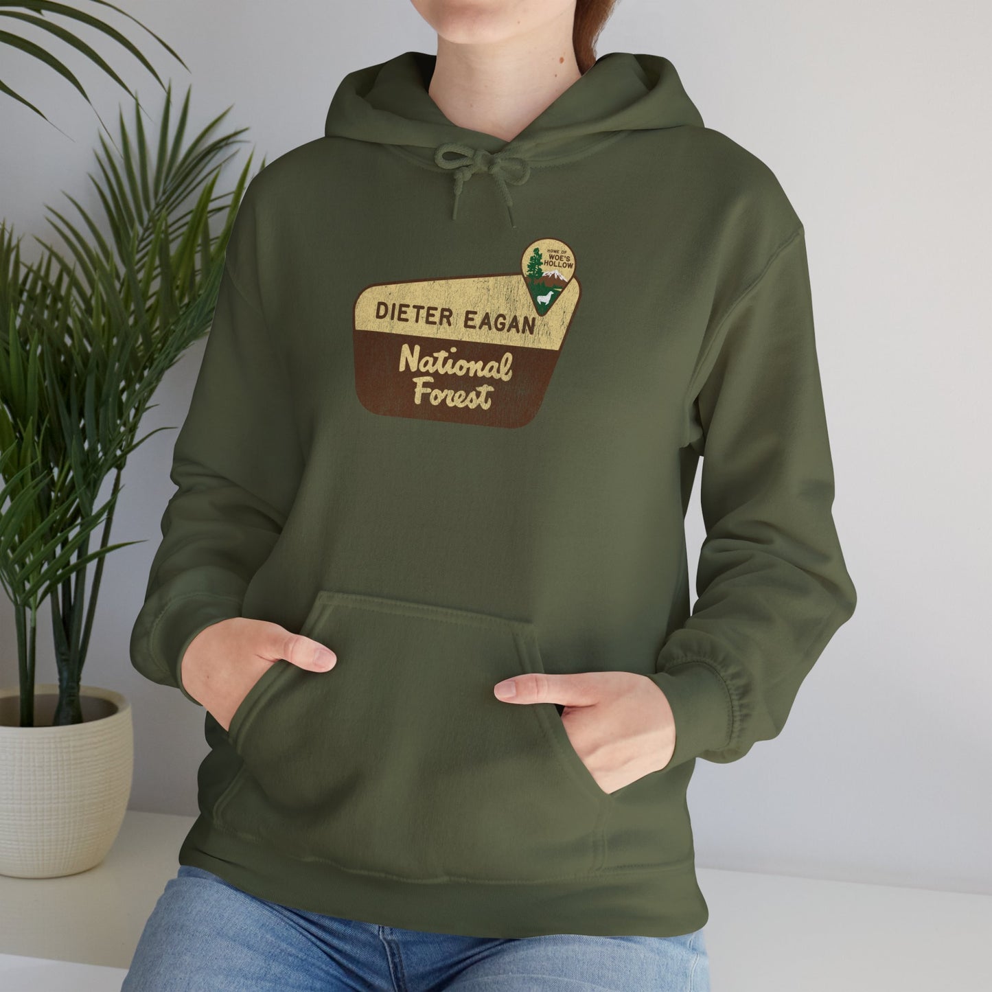 Female model wearing a military green colored Soft Unisex Hoodie Heavy Blend Hooded Sweatshirt featuring the logo graphic of the Dieter Eagan National Forest inspired by the TV show Severance in a vintage distressed ink-stamped worn-in style.