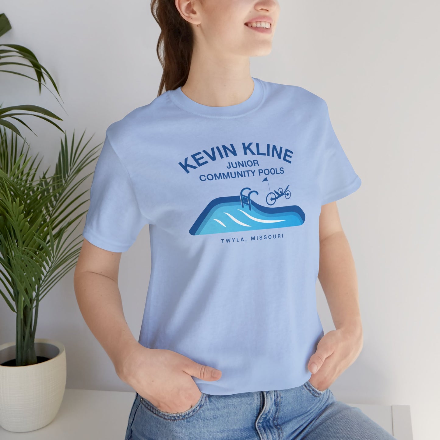 Woman wearing light blue DTF St. Louis Kevin Kline Junior Community Pools graphic tee with pool and recumbent trike.