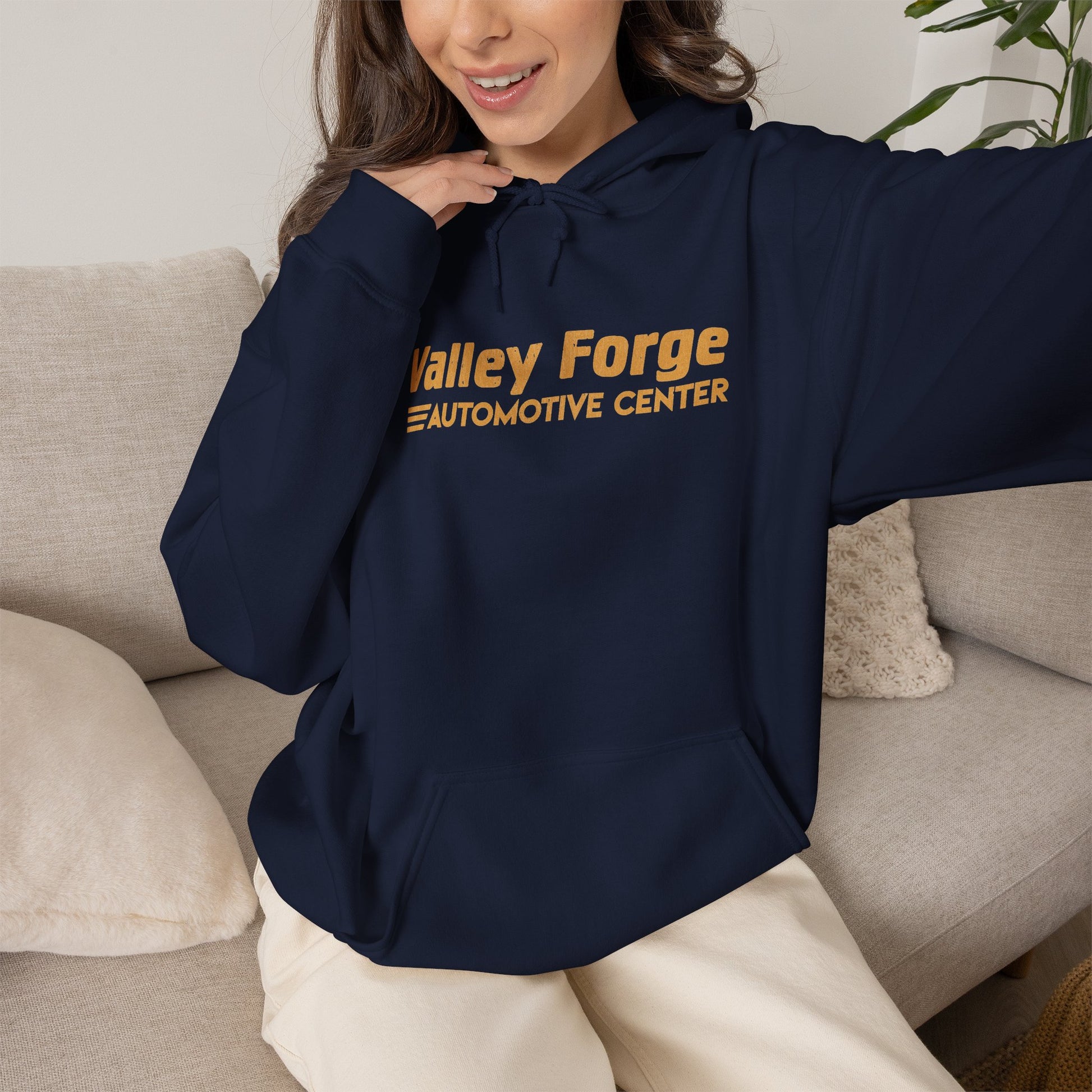 A woman wearing a navy blue hoodie with gold text reading 'Valley Forge Automotive Center' sits on a light beige couch in a home setting.