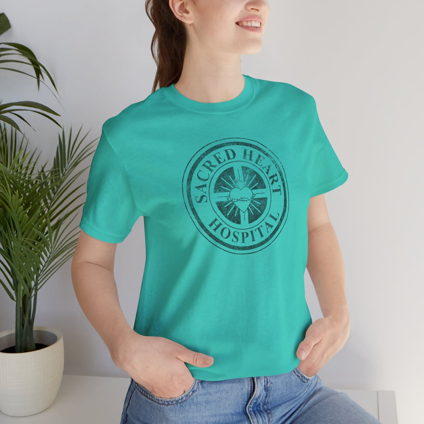 Woman wearing teal blue Bella Canvas 3001 tee with vintage distressed Sacred Heart Hospital logo from Scrubs printed on chest.