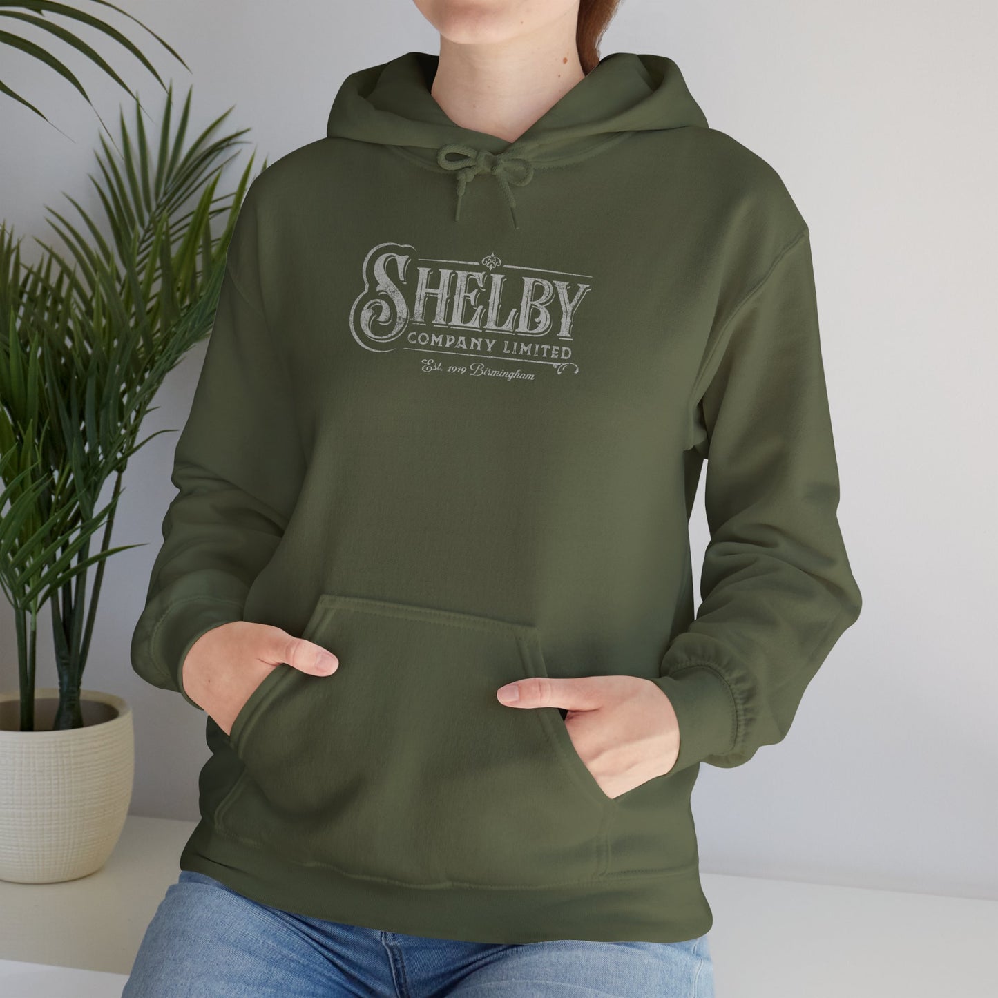 Front view of a Female model wearing a military green Soft Unisex Hoodie Heavy Blend Hooded Sweatshirt featuring the fictional logo Shelby Company Limited from the TV series Peaky Blinders in a vintage distressed style.