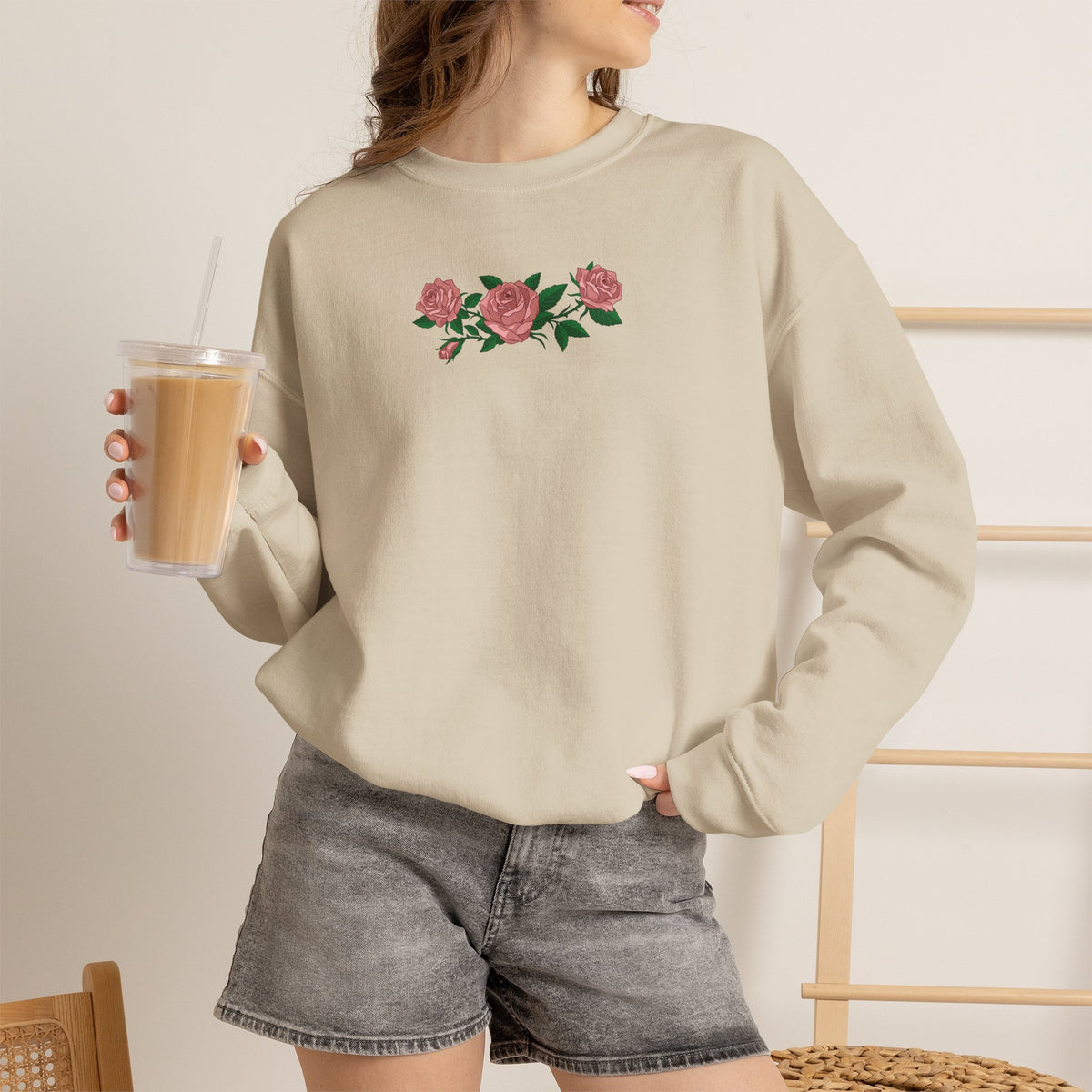 Person wearing a beige tan crewneck sweatshirt with three pink roses and green leaves printed across the chest, inspired by Ruth of Widow's Bay.