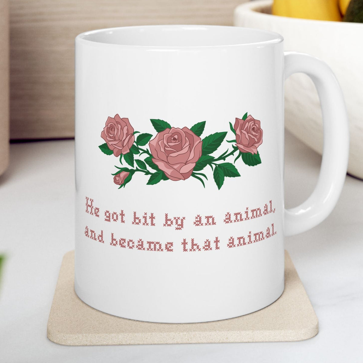 white ceramic mug with a printed design of pink roses and green leaves, featuring the text 'He got bit by an animal, and became that animal' in cross stitch style, inspired by Ruth on Widows Bay. On a beige coaster on a light-colored surface.