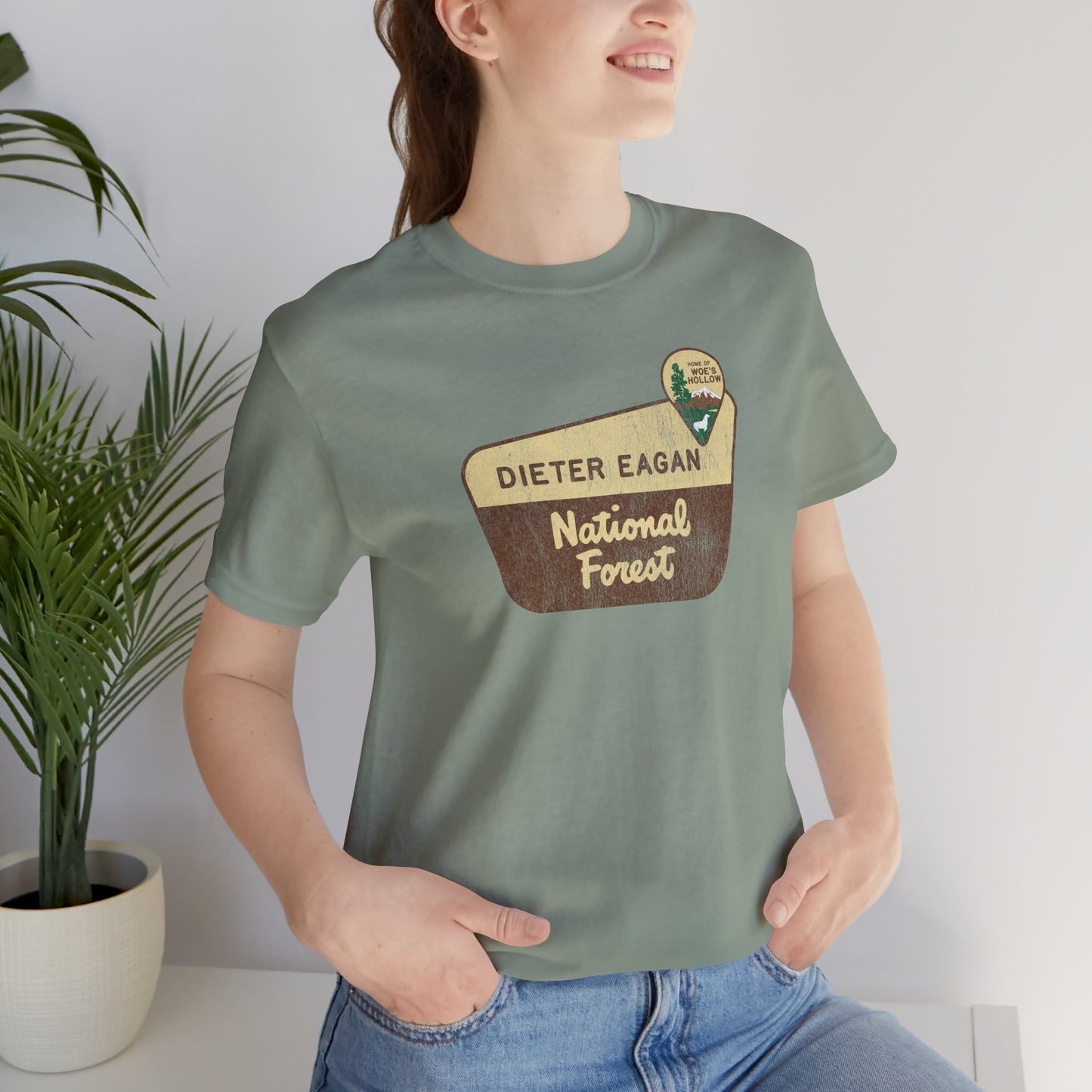 Female model wearing a sage green colored soft Bella+Canvas short-sleeve t-shirt with the logo graphic of the Dieter Eagan National Forest inspired by the TV show Severance in a vintage distressed ink-stamped worn-in style.