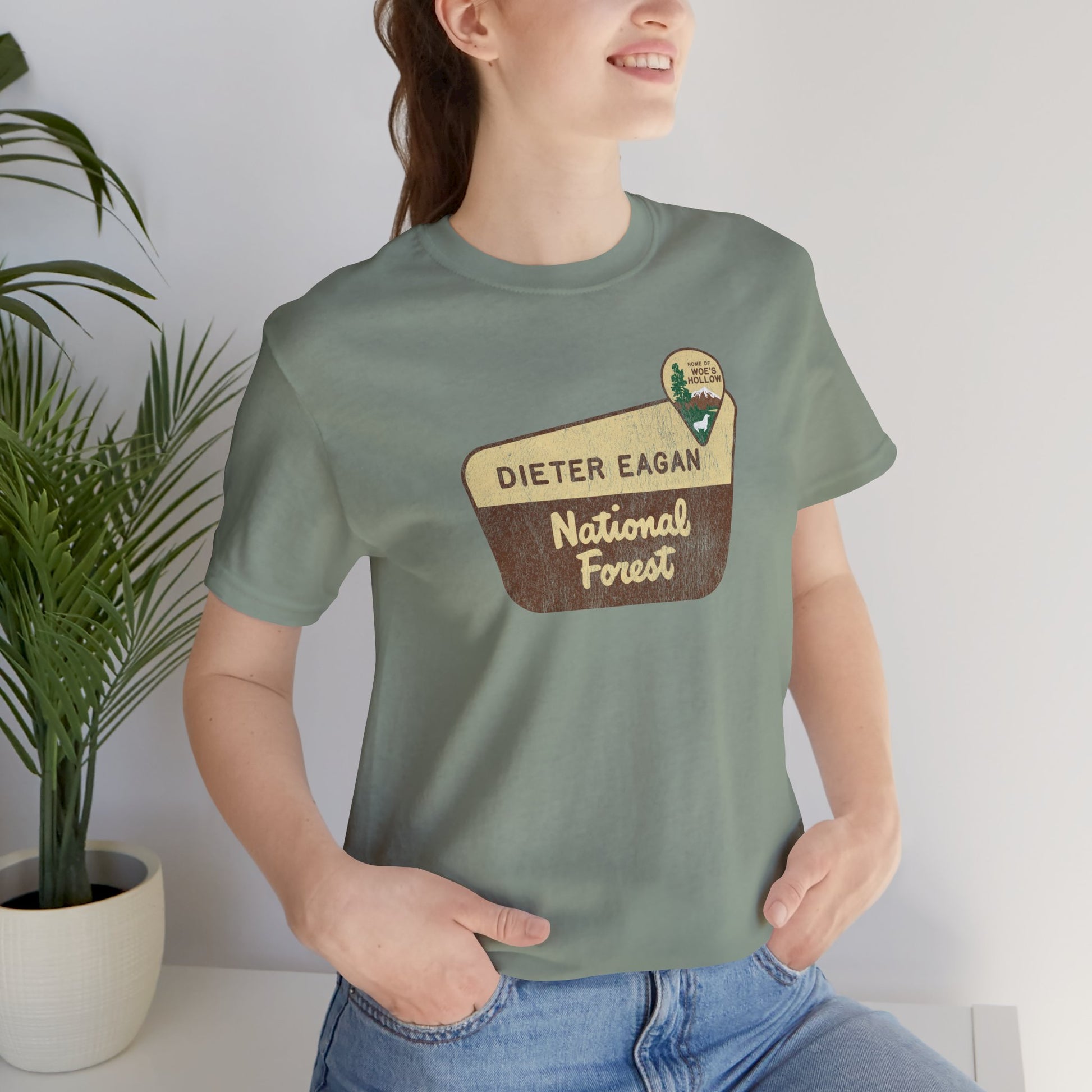 Female model wearing a sage green colored soft Bella+Canvas short-sleeve t-shirt with the logo graphic of the Dieter Eagan National Forest inspired by the TV show Severance in a vintage distressed ink-stamped worn-in style.