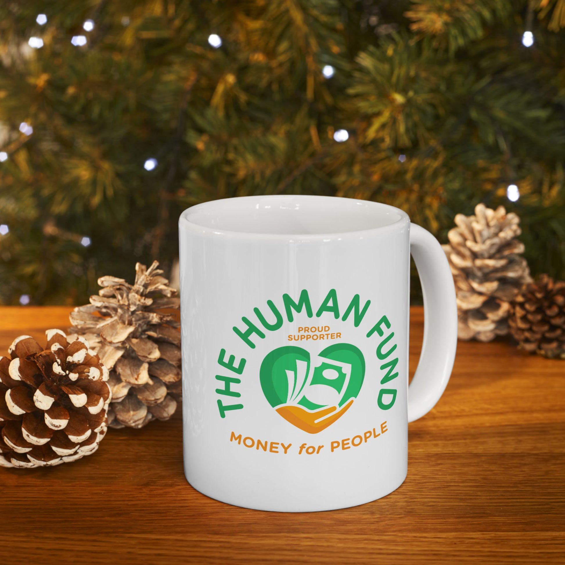White mug with 'The Human Fund' logo on a wooden surface with pinecones and a Christmas tree in the background.
