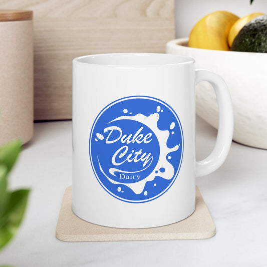 White ceramic mug with the blue Duke City Dairy logo printed on both sides, inspired by the TV show Pluribus, set in a kitchen.