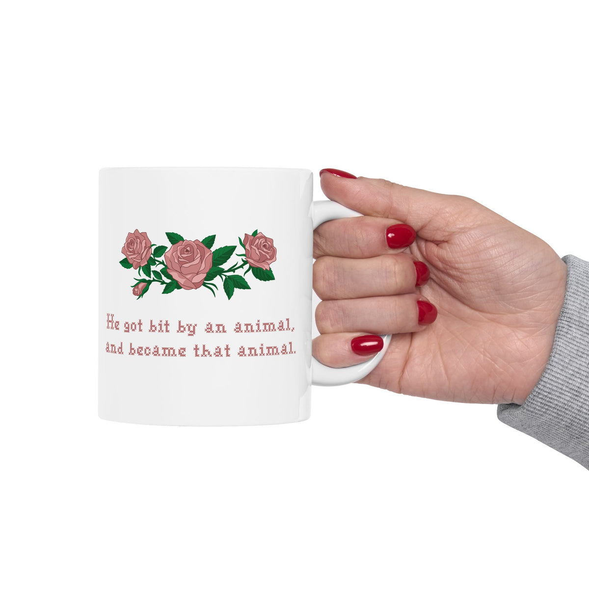 Woman holding an 11oz white ceramic mug with a printed design of pink roses and green leaves, featuring the text 'He got bit by an animal, and became that animal' in cross stitch style, inspired by Ruth on Widows Bay