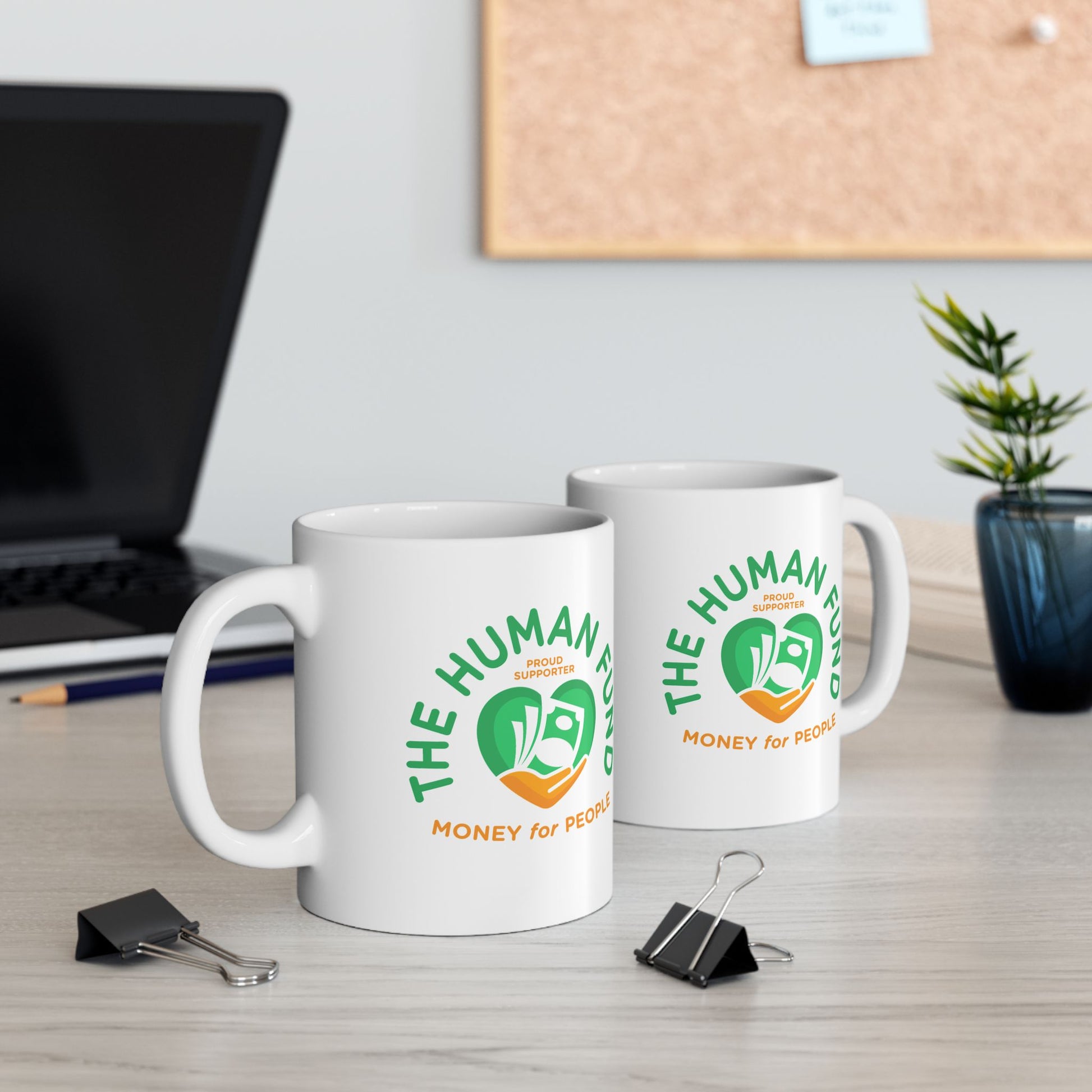 Two white mugs with 'The Human Fund' logo on a desk with office items.