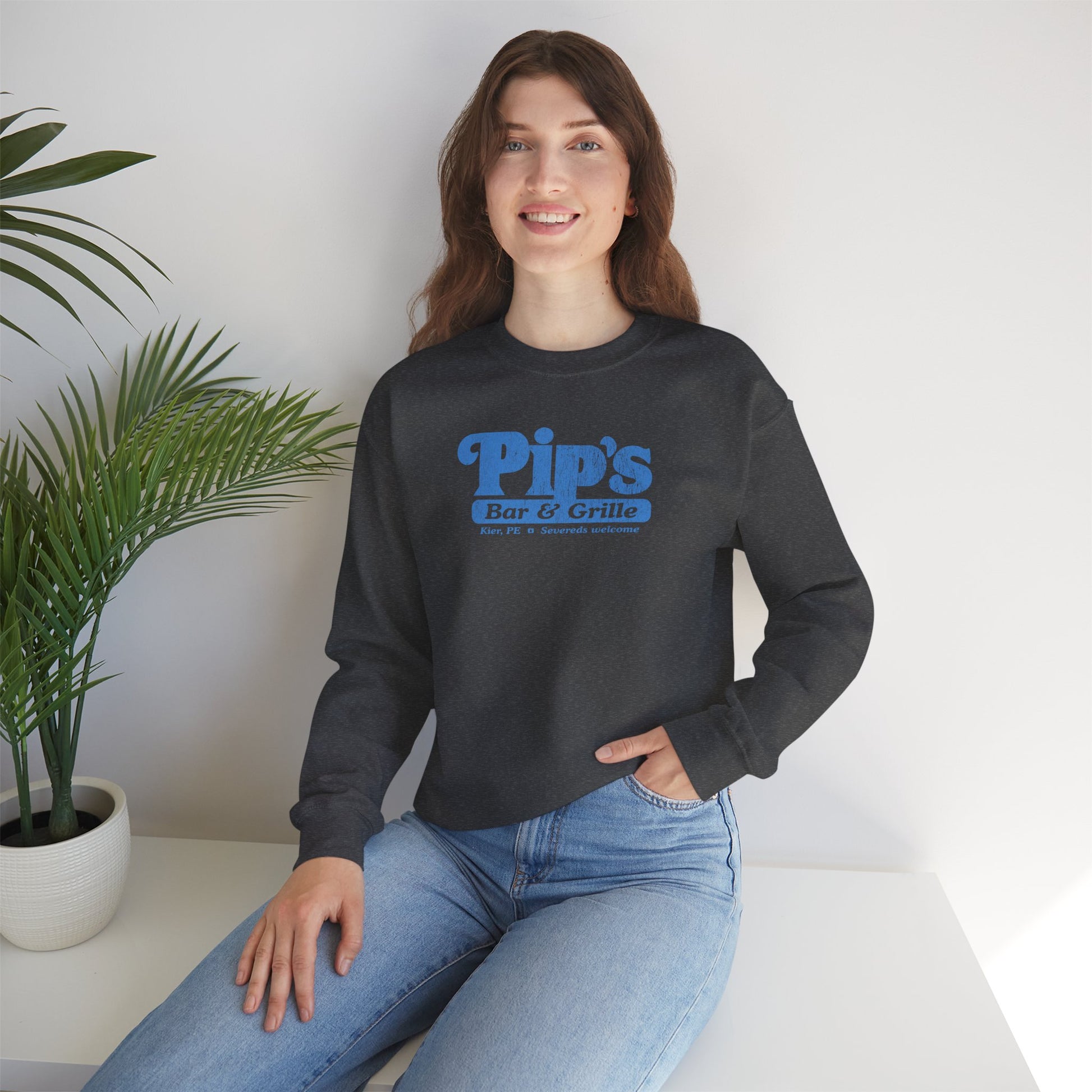 Female model wearing a dark heather gray soft Gildan 18000 sweatshirt featuring the Pips Bar and Grill logo Severeds Welcome from the TV show Severance in a vintage distressed ink-stamped worn-in style.