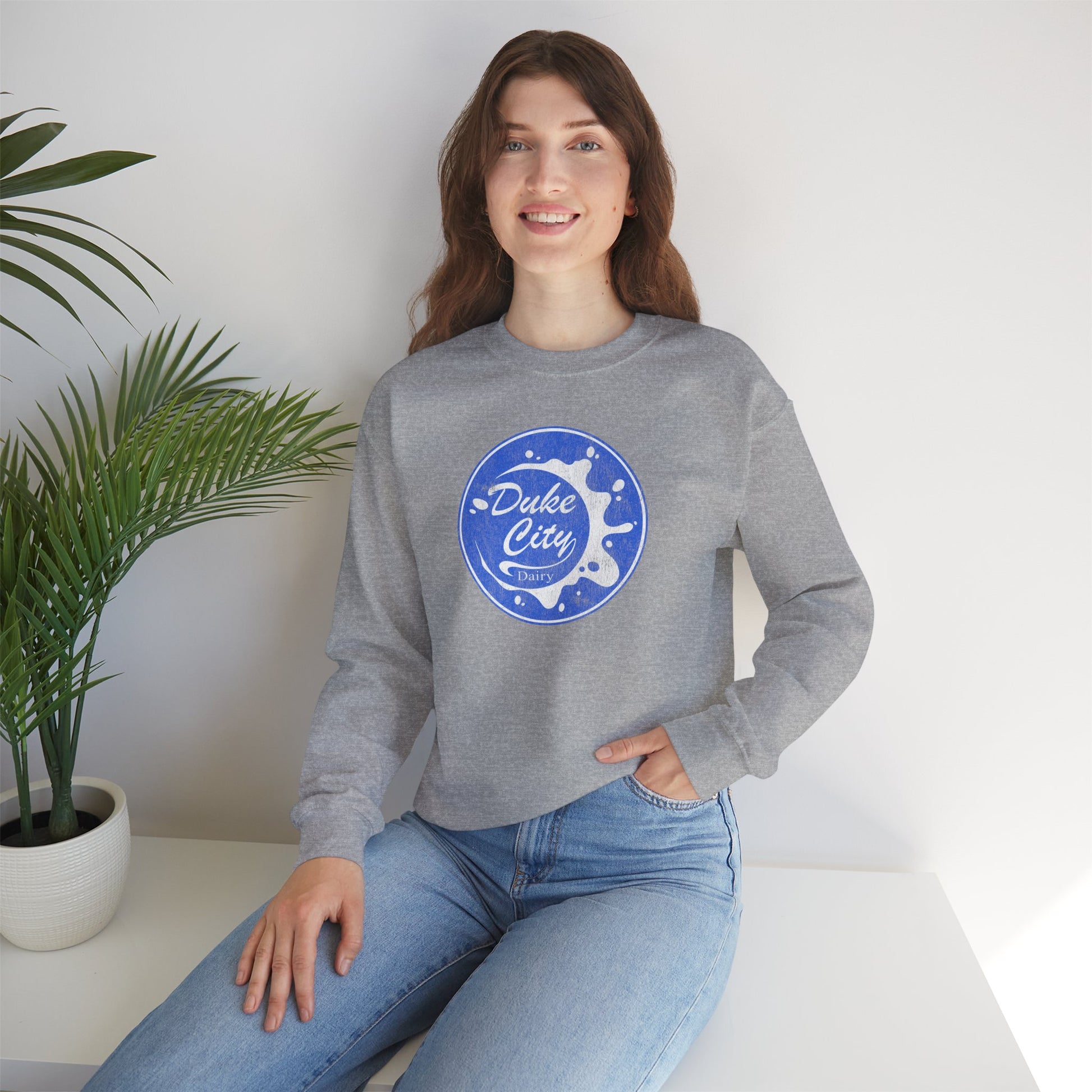 Woman wearing a heather gray sweatshirt with 'Duke City' logo design from Pluribus, sitting on a white surface with a plant in the background.