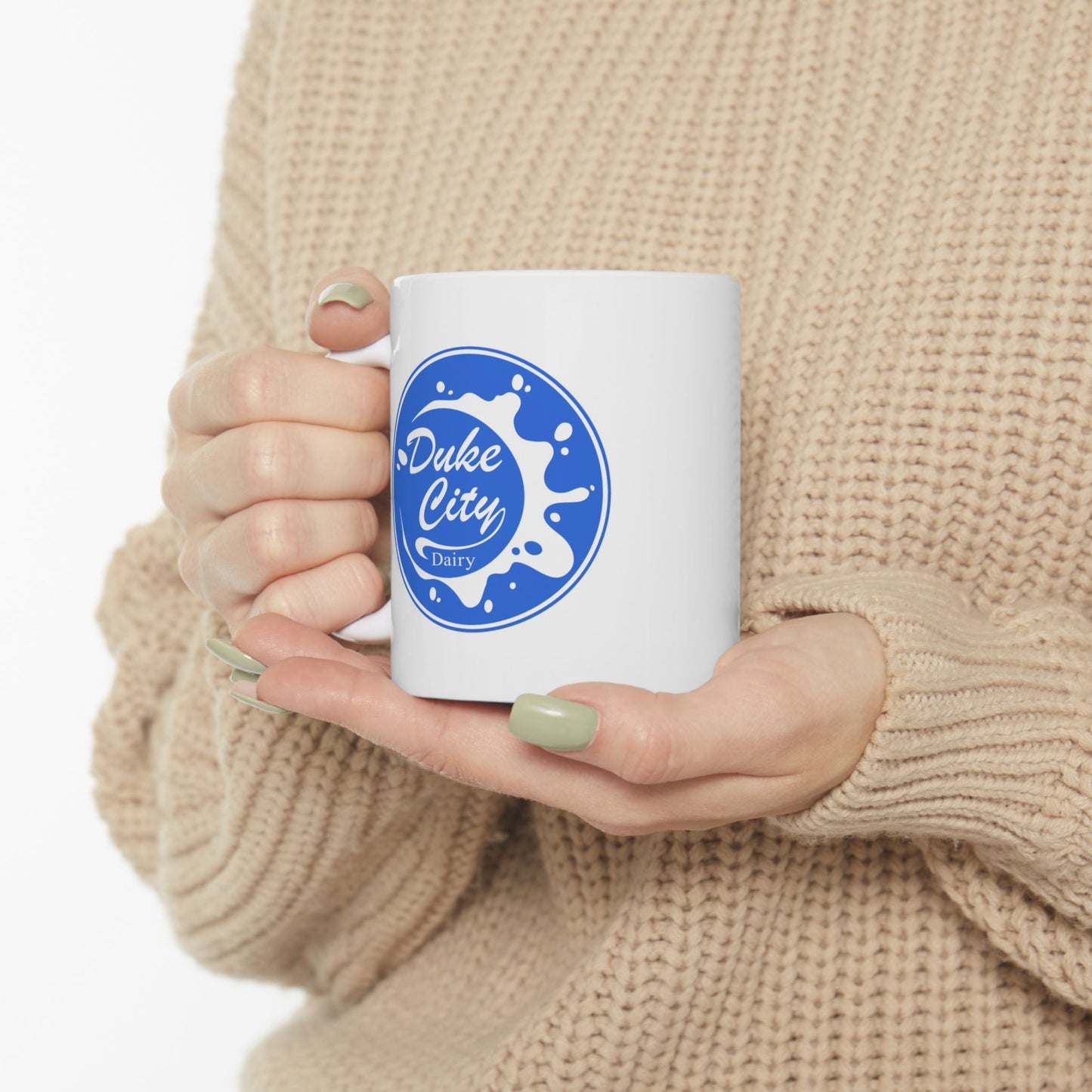 Person holding a white mug with a blue 'Duke City Dairy' logo from Pluribus, wearing a beige sweater.