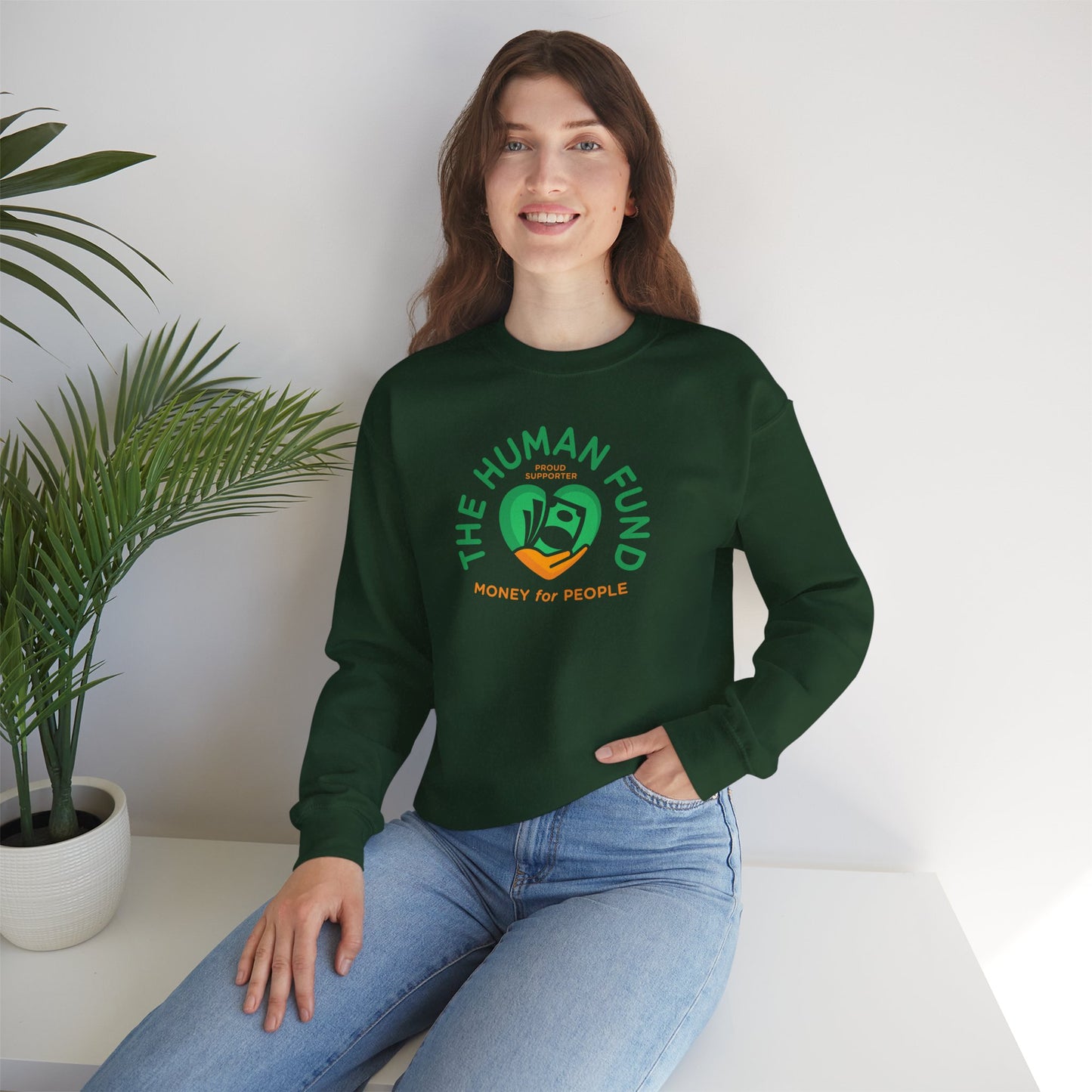 Person wearing a forest green sweatshirt with 'The Human Fund' logo sitting on a white surface with a plant in the background.