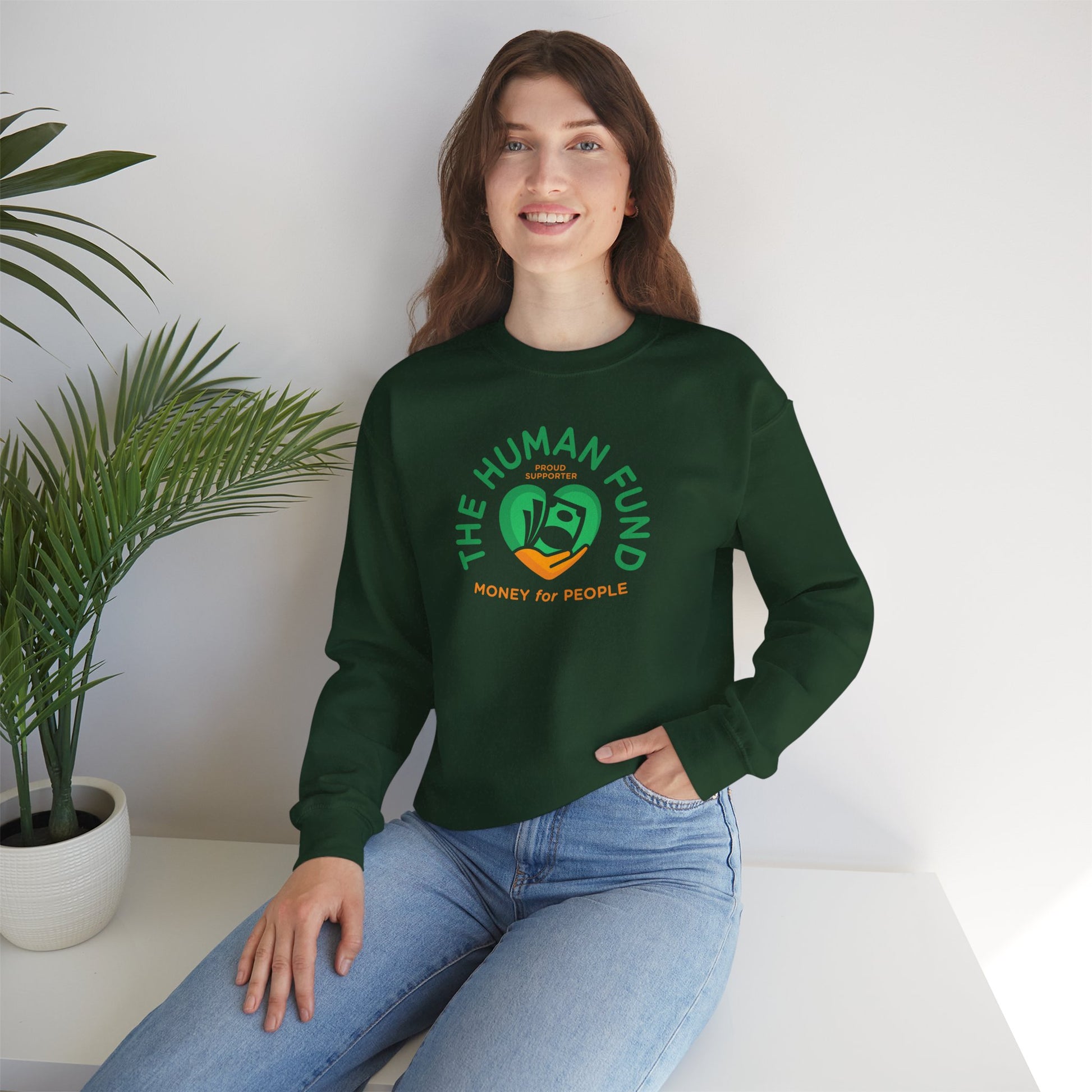 Person wearing a forest green sweatshirt with 'The Human Fund' logo sitting on a white surface with a plant in the background.