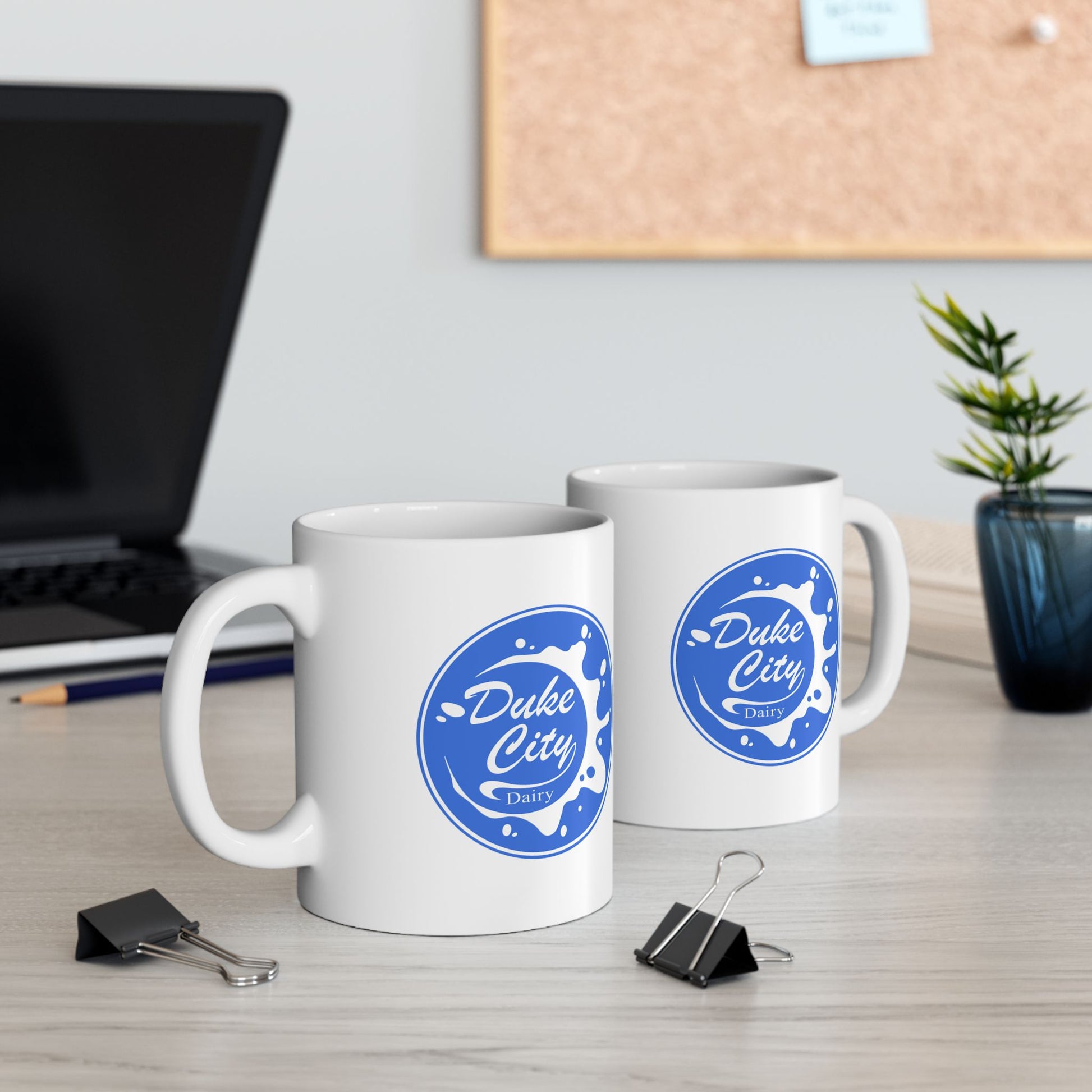 Two white mugs with blue 'Duke City' logo on a desk with office supplies and a plant.