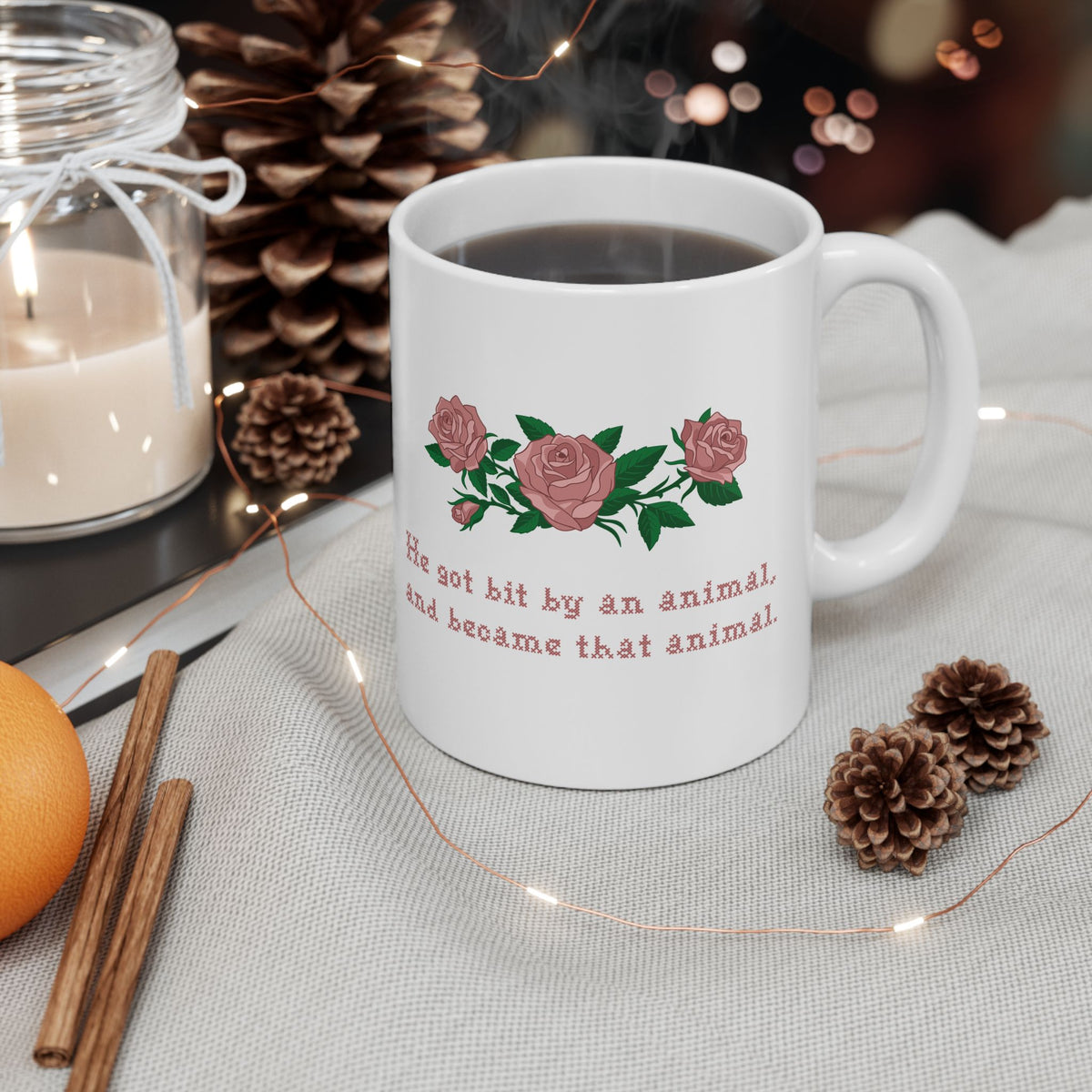 White ceramic mug filled with coffee with a printed design of pink roses and green leaves, featuring the text 'He got bit by an animal, and became that animal' in cross stitch style, inspired by Ruth on Widows Bay
