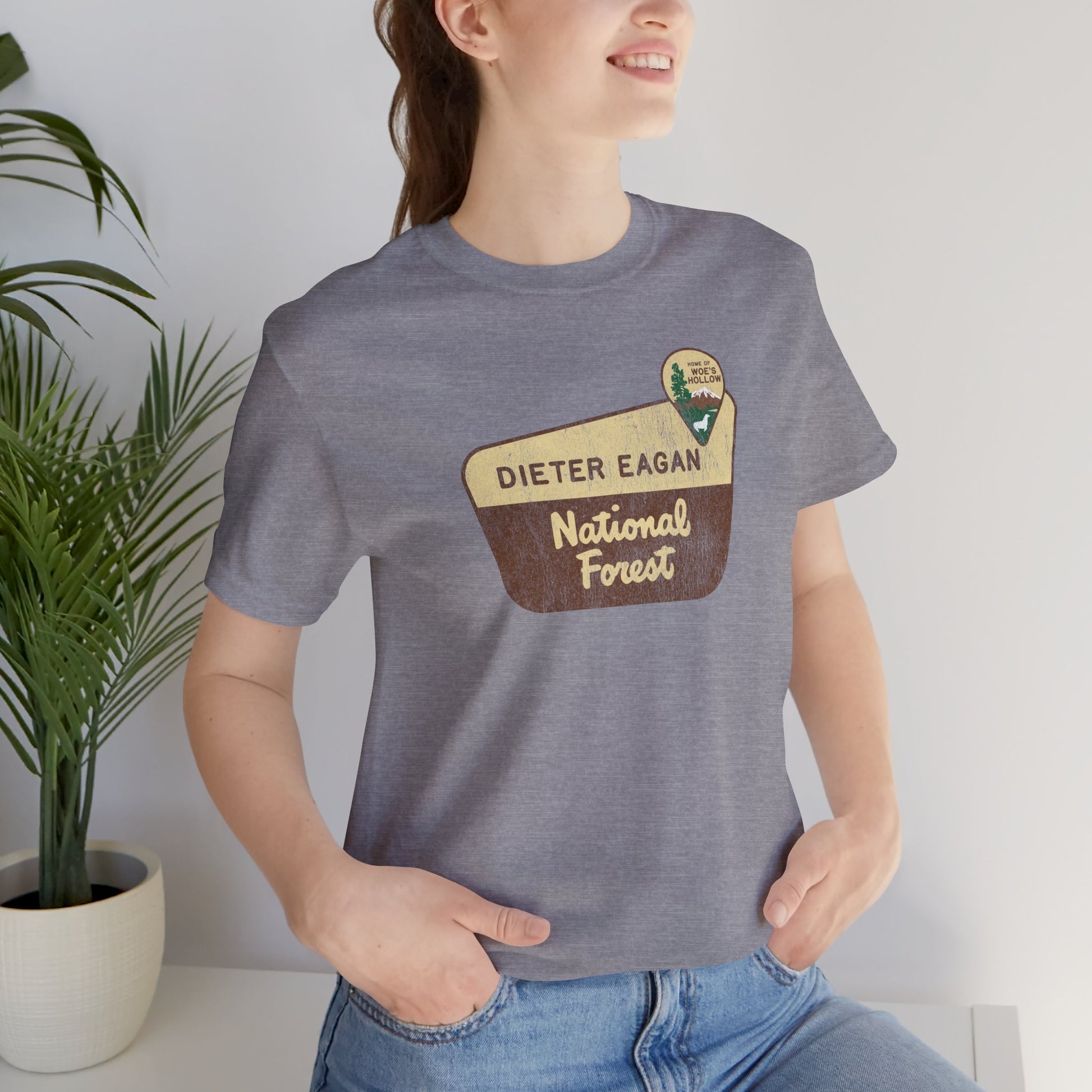 Female model wearing a heather storm colored soft Bella+Canvas short-sleeve t-shirt with the logo graphic of the Dieter Eagan National Forest inspired by the TV show Severance in a vintage distressed ink-stamped worn-in style.