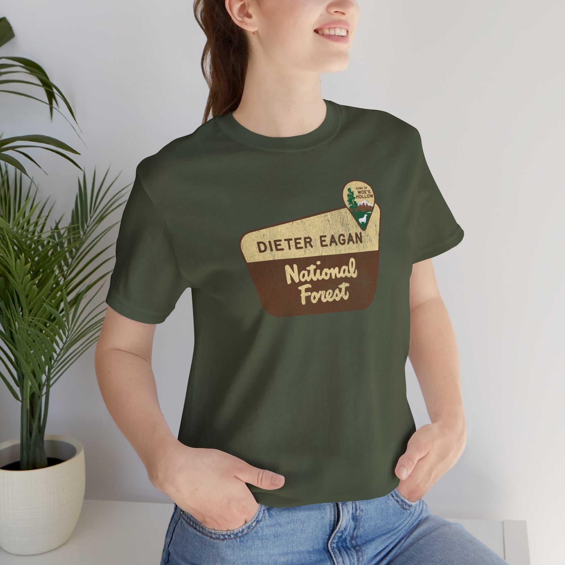 Female model wearing a military green colored soft Bella+Canvas short-sleeve t-shirt with the logo graphic of the Dieter Eagan National Forest inspired by the TV show Severance in a vintage distressed ink-stamped worn-in style.