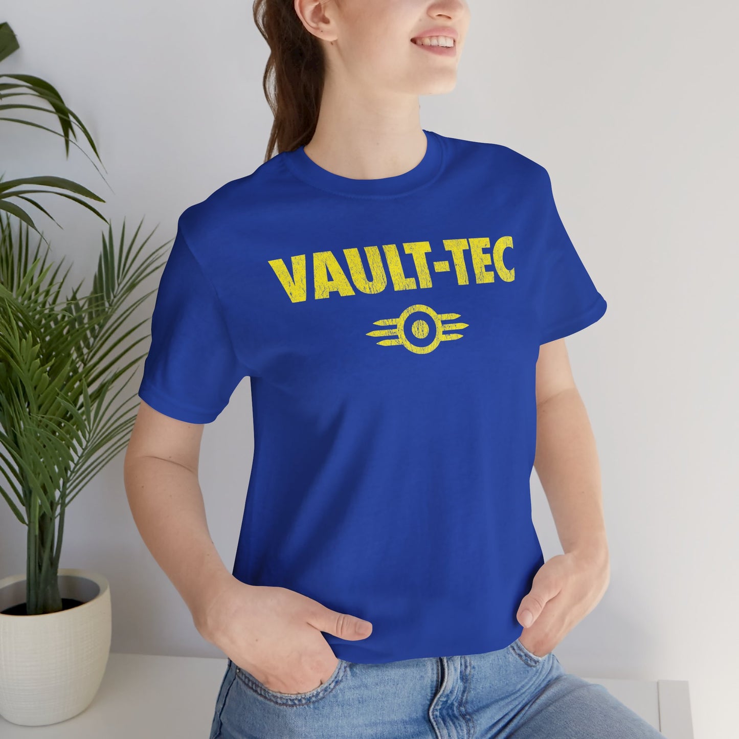 Woman wearing a royal blue t-shirt featuring the Vault-Tec logo graphic from the video game Fallout in a vintage distressed style. 