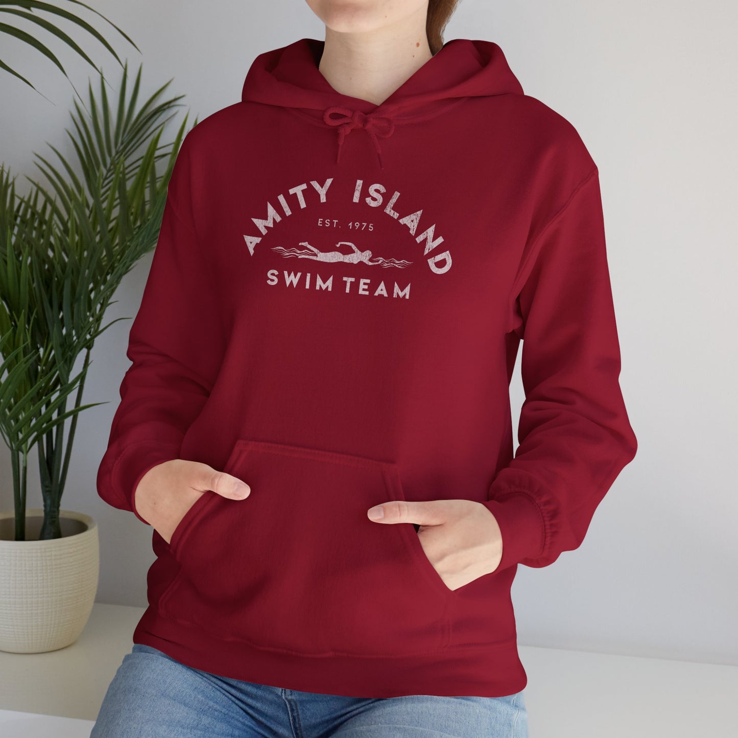 Female model wearing a lifeguard cardinal red colored Soft Unisex Hoodie Heavy Blend Hooded Sweatshirt featuring an Amity Island Swim Team logo graphic, designed by ScriptedBrand, and  inspired by the movie Jaws in a vintage distressed style.