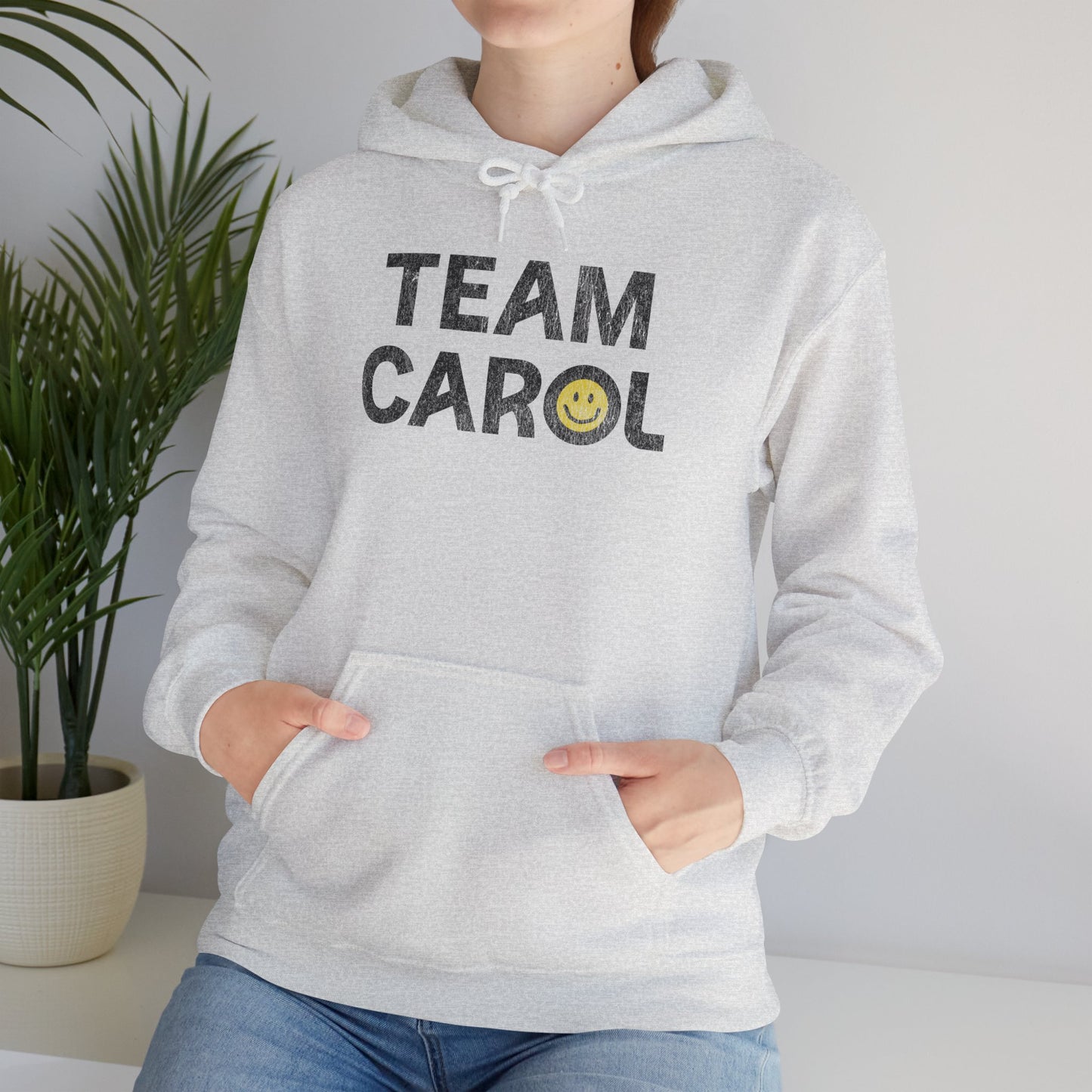 Woman wearing ash unisex Heavy Blend pullover hoodie sweatshirt with TEAM CAROL vintage distressed smiley graphic