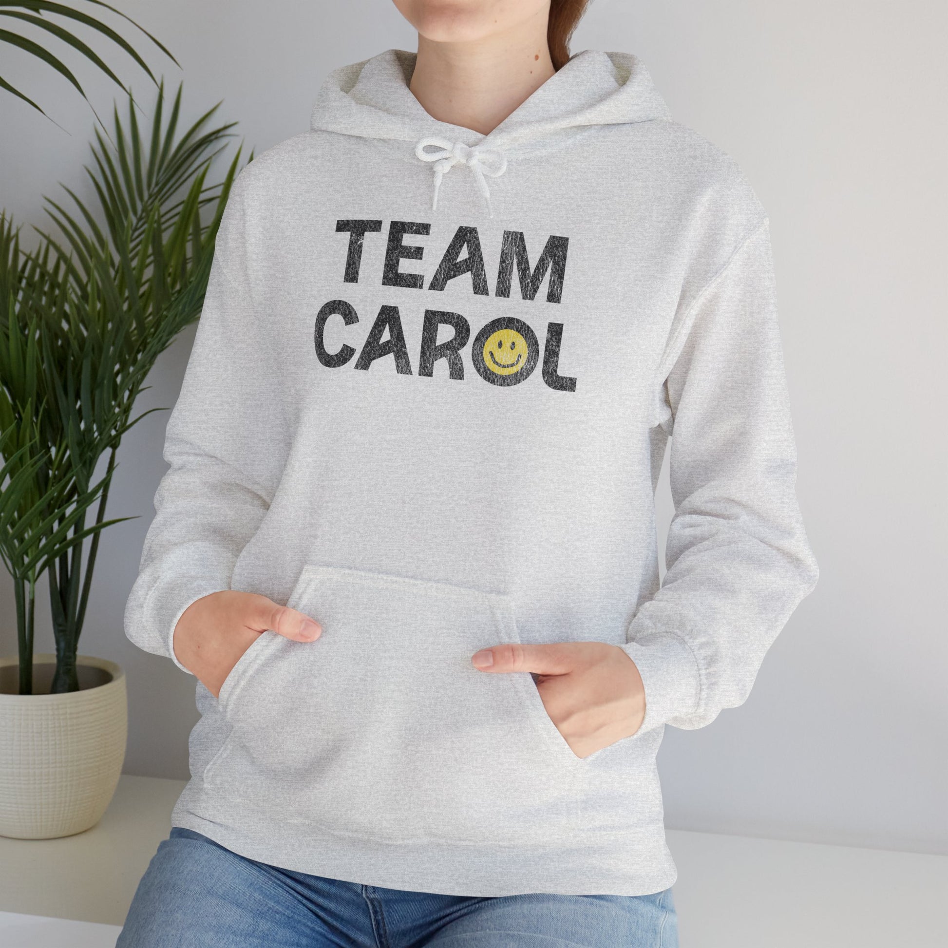 Woman wearing ash unisex Heavy Blend pullover hoodie sweatshirt with TEAM CAROL vintage distressed smiley graphic