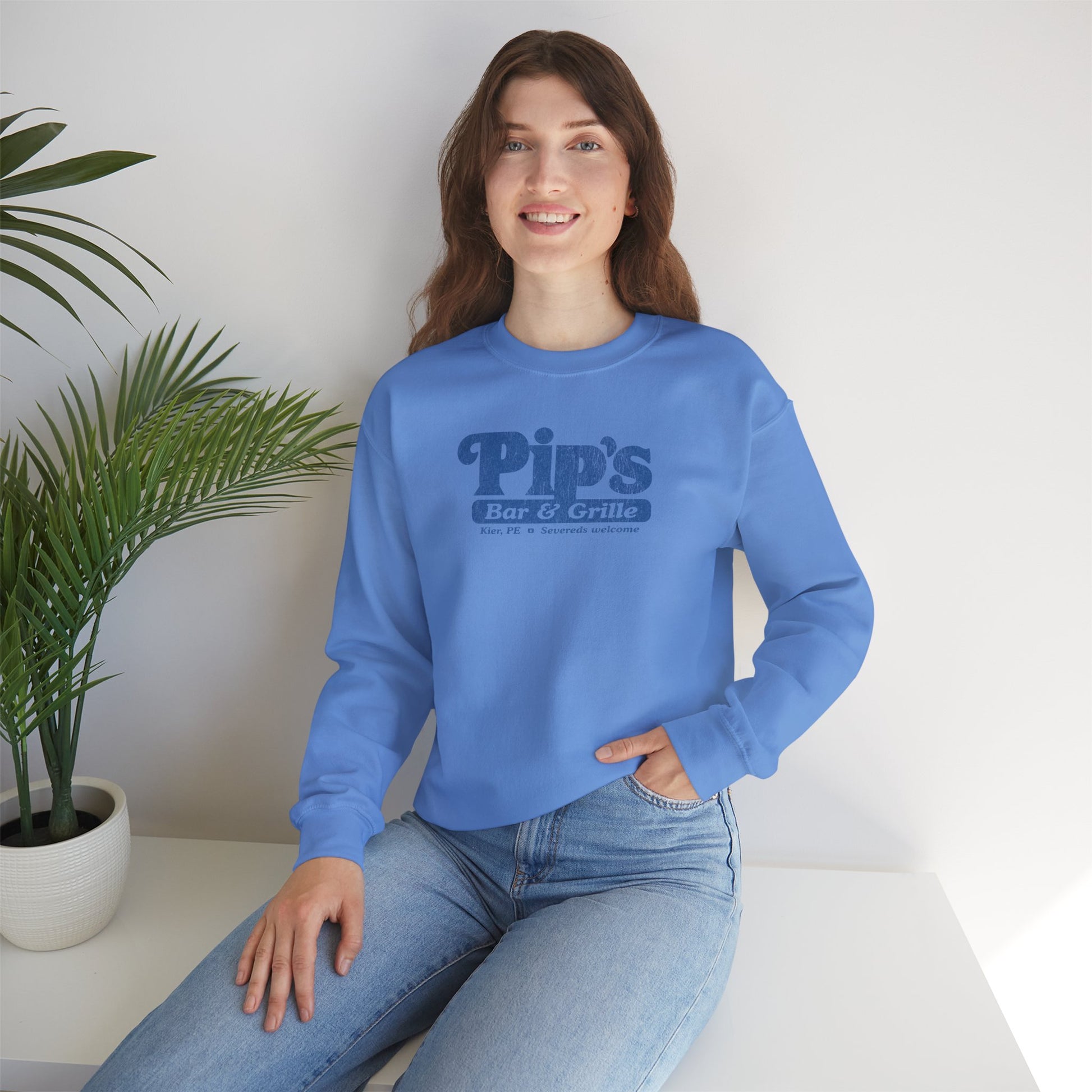 Female model wearing a Carolina blue Soft Gildan 18000 sweatshirt featuring the Pips Bar and Grill logo Severeds Welcome from the TV show Severance in a vintage distressed ink-stamped worn-in style.