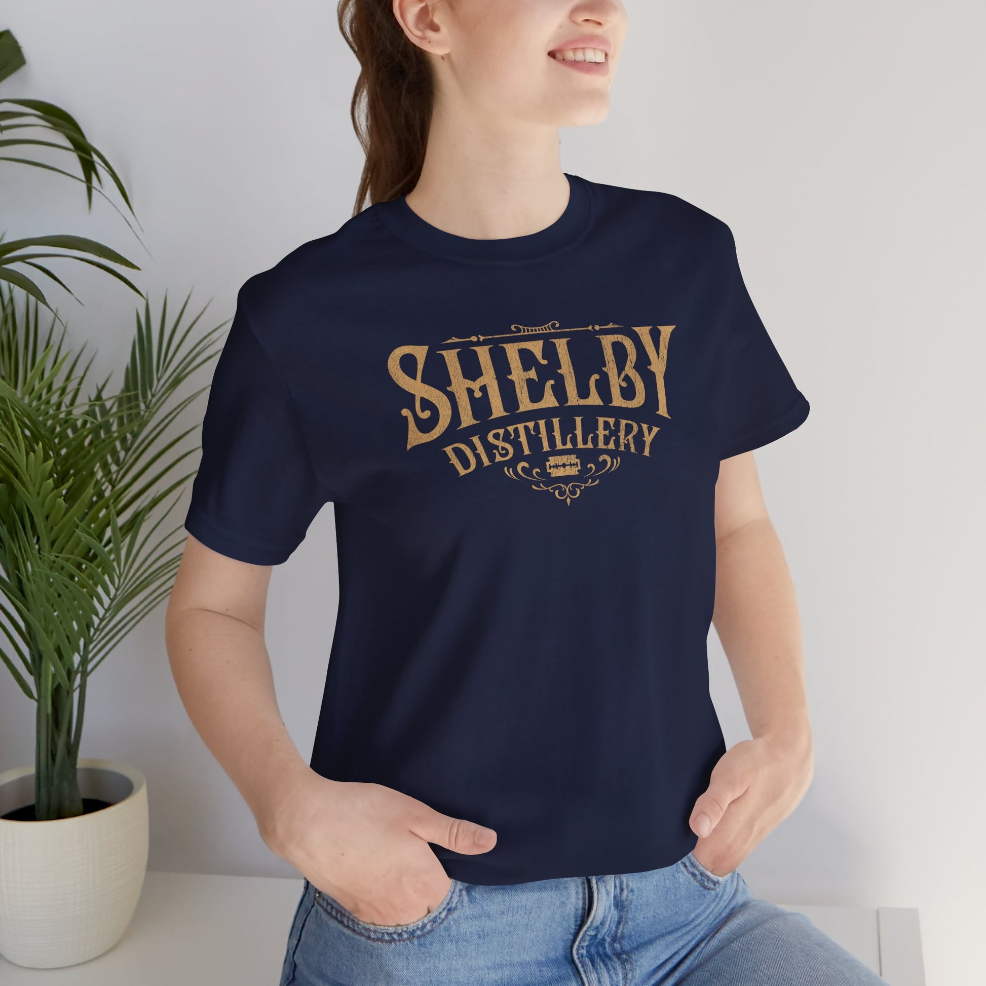 Female model wearing a navy blue Soft Bella+Canvas 3001 short-sleeve t-shirt featuring the Shelby Company Limited Distillery Logo Graphic inspired by the TV show and movie Peaky Blinders in a vintage distressed style, sold by ScriptedBrand.
