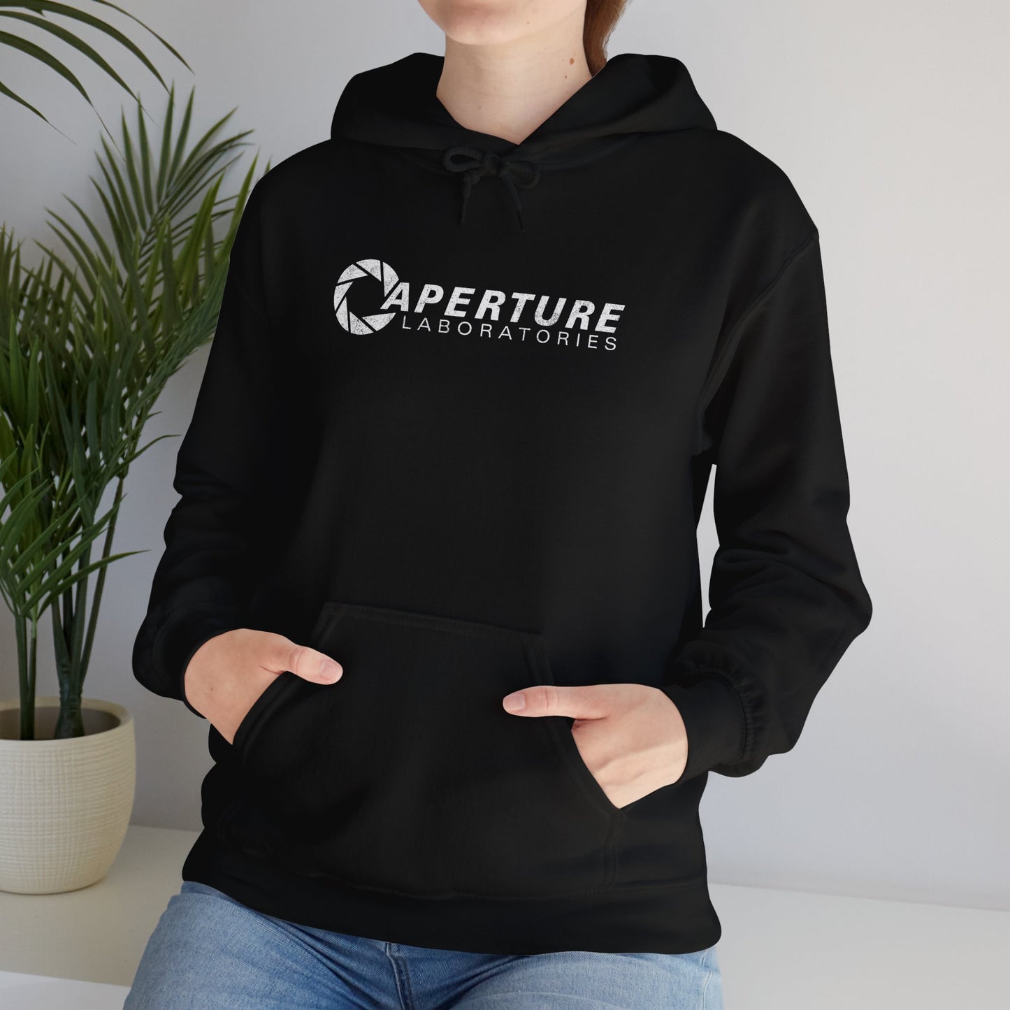 Female model wearing a black colored Unisex Hoodie Heavy Blend Hooded Sweatshirt featuring the Aperture Laboratories logo from the video game Portal and Portal 2 in a vintage distressed style, sold by ScriptedBrand.