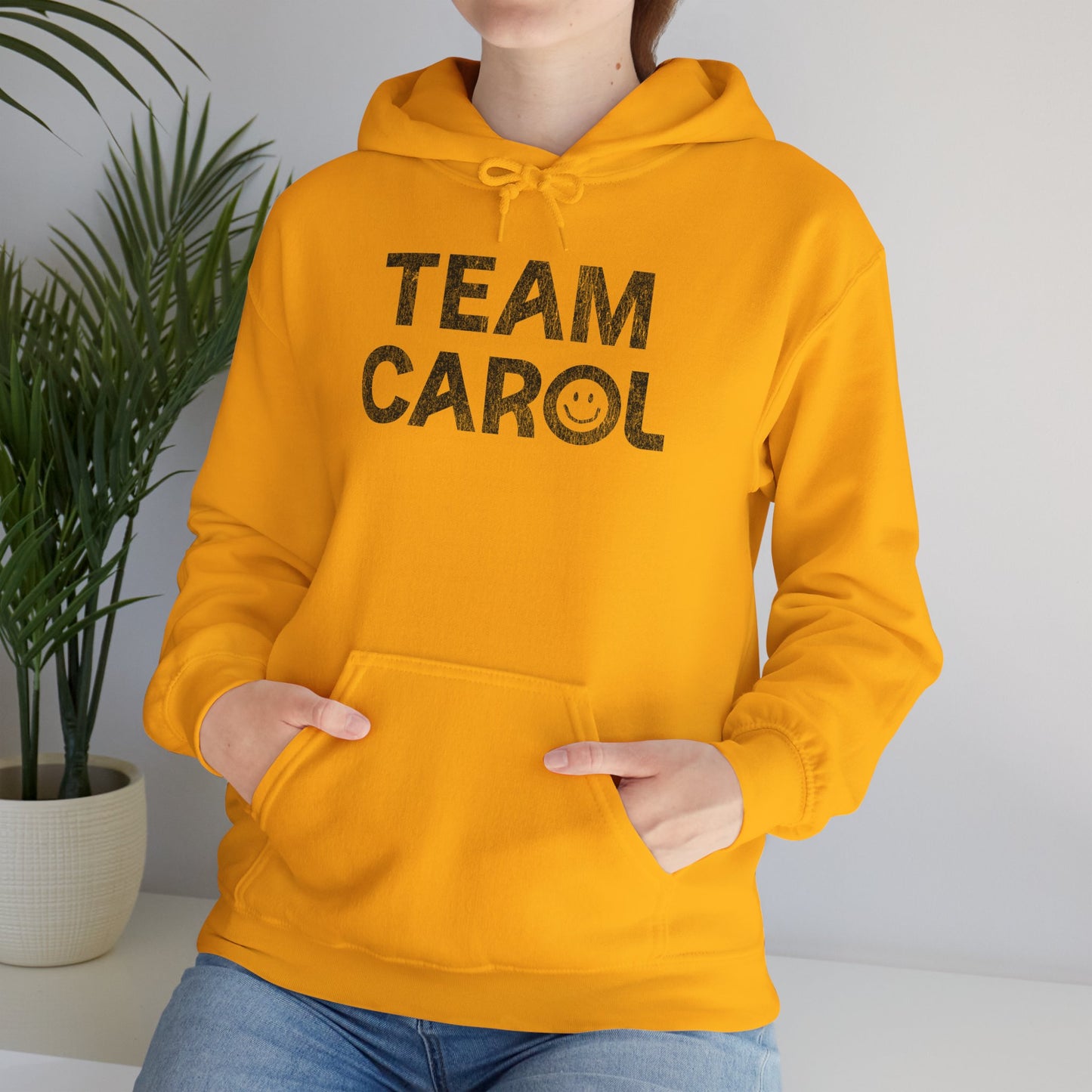Woman wearing gold yellow unisex Heavy Blend pullover hoodie sweatshirt with TEAM CAROL vintage distressed smiley graphic
