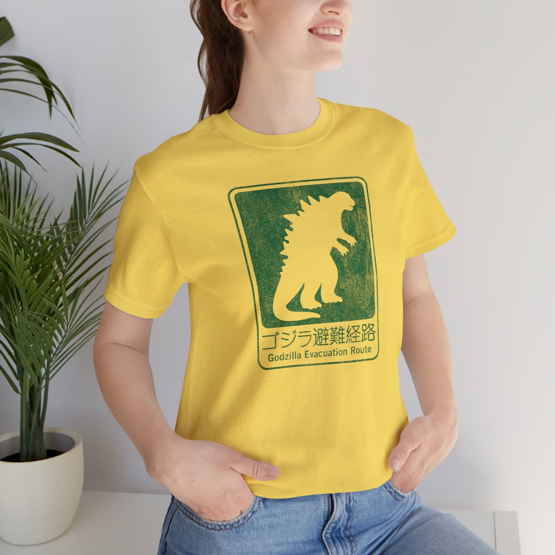 Woman wearing yellow and green Godzilla Evacuation Route distressed graphic t-shirt inspired by Monarch Legacy of Monsters, soft Bella+Canvas tee