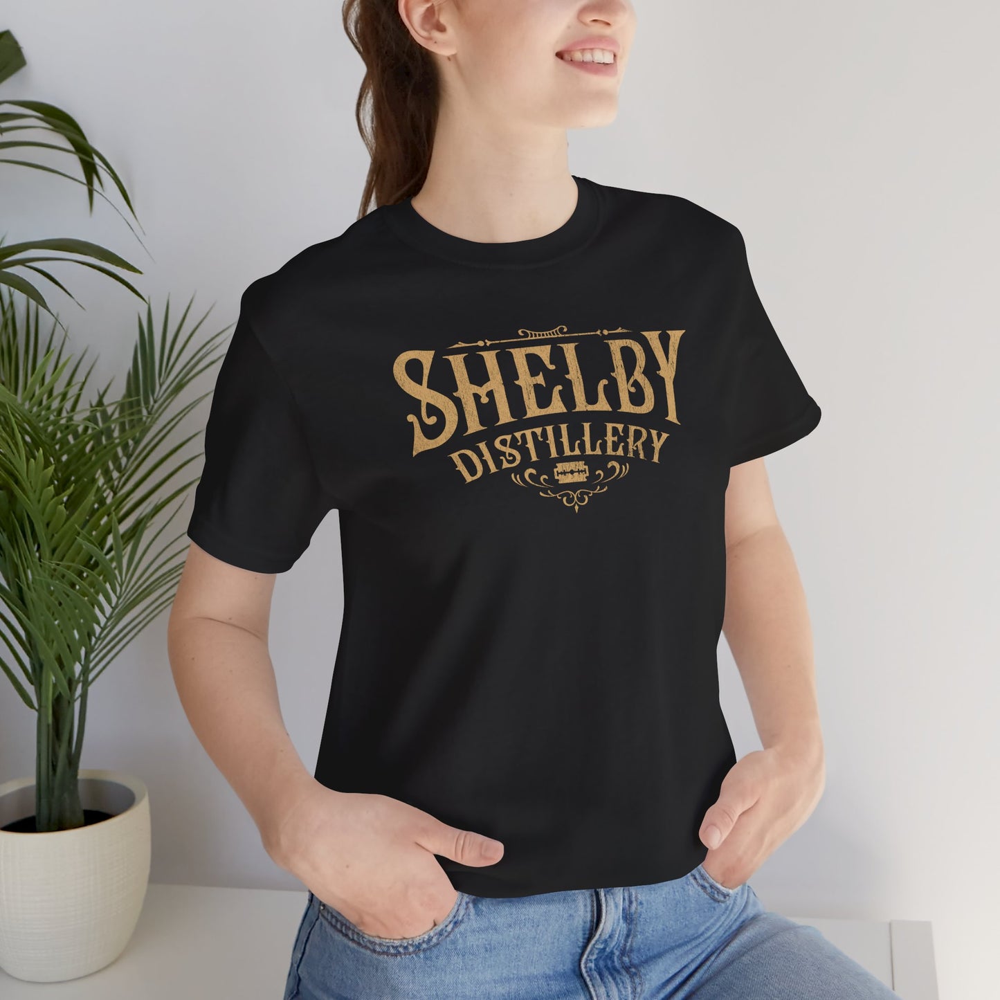 Female model wearing a black Soft Bella+Canvas 3001 short-sleeve t-shirt featuring the Shelby Company Limited Distillery Logo Graphic inspired by the TV show and movie Peaky Blinders in a vintage distressed style, sold by ScriptedBrand.