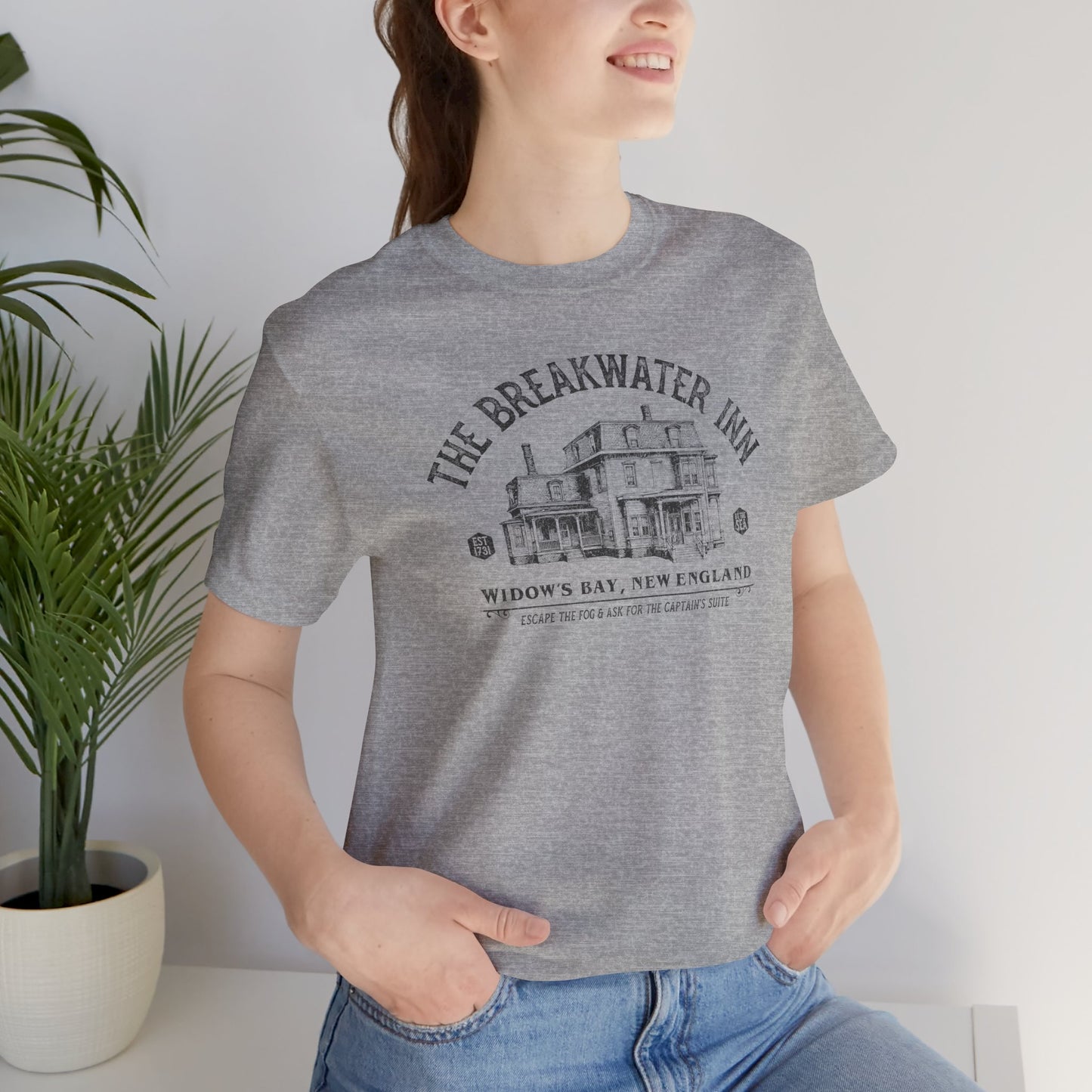 Woman wearing Heather gray 'The Breakwater Inn' vintage illustrated t-shirt inspired by Widow's Bay Apple TV series, soft Bella+Canvas cotton fan tee