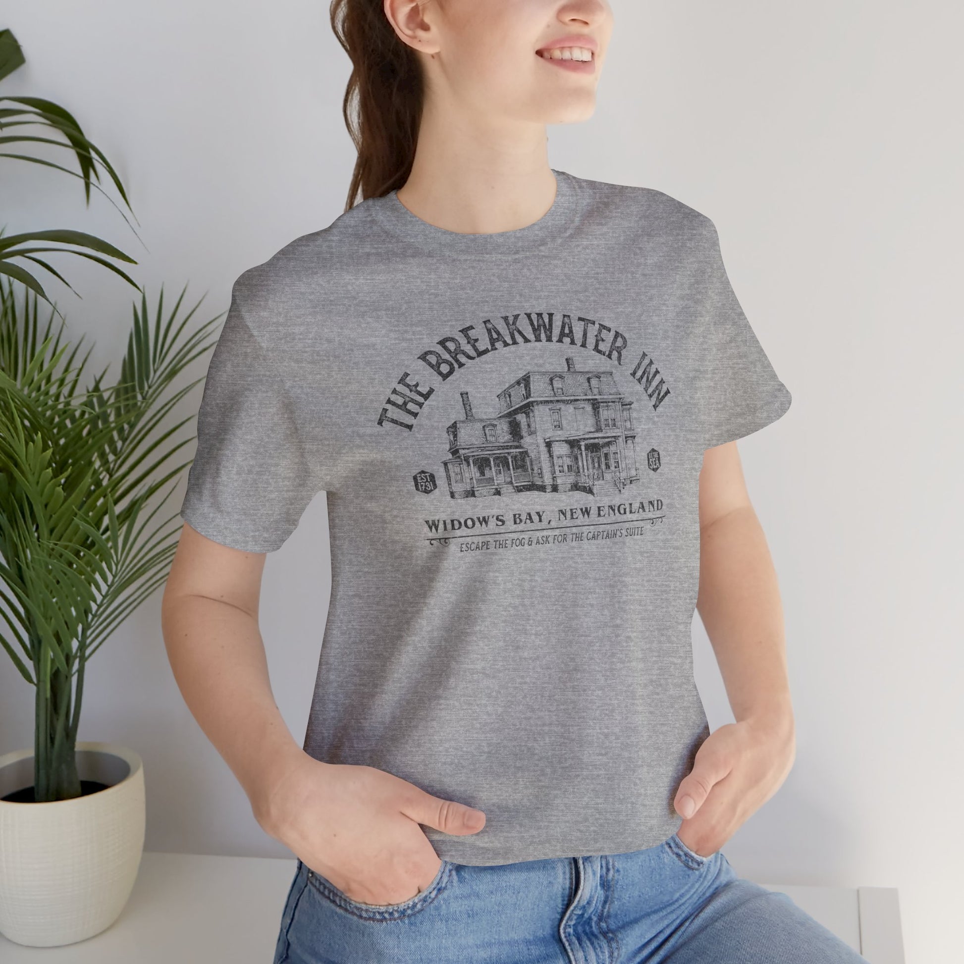 Woman wearing Heather gray 'The Breakwater Inn' vintage illustrated t-shirt inspired by Widow's Bay Apple TV series, soft Bella+Canvas cotton fan tee