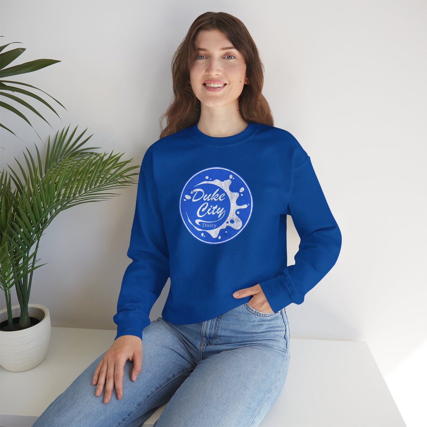 Woman wearing a royal blue sweatshirt with 'Duke City' logo design from Pluribus, sitting on a white surface with a plant in the background.