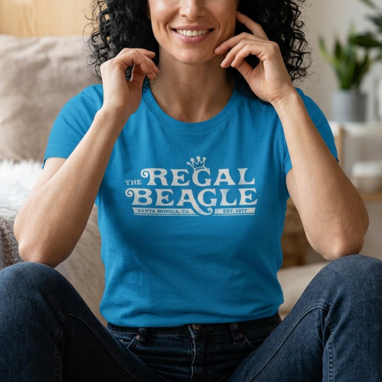 A smiling woman with curly dark hair sits cross-legged indoors wearing a bright blue graphic T-shirt that reads “THE REGAL BEAGLE” in white lettering, with dark jeans and a softly lit home interior in the background.