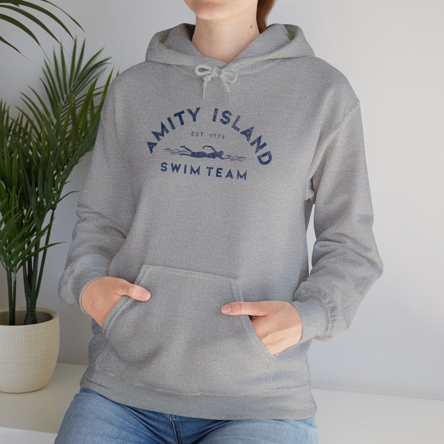 Female model wearing a sport heather grey colored Soft Unisex Hoodie Heavy Blend Hooded Sweatshirt featuring an Amity Island Swim Team logo graphic, designed by ScriptedBrand, and  inspired by the movie Jaws in a vintage distressed style.