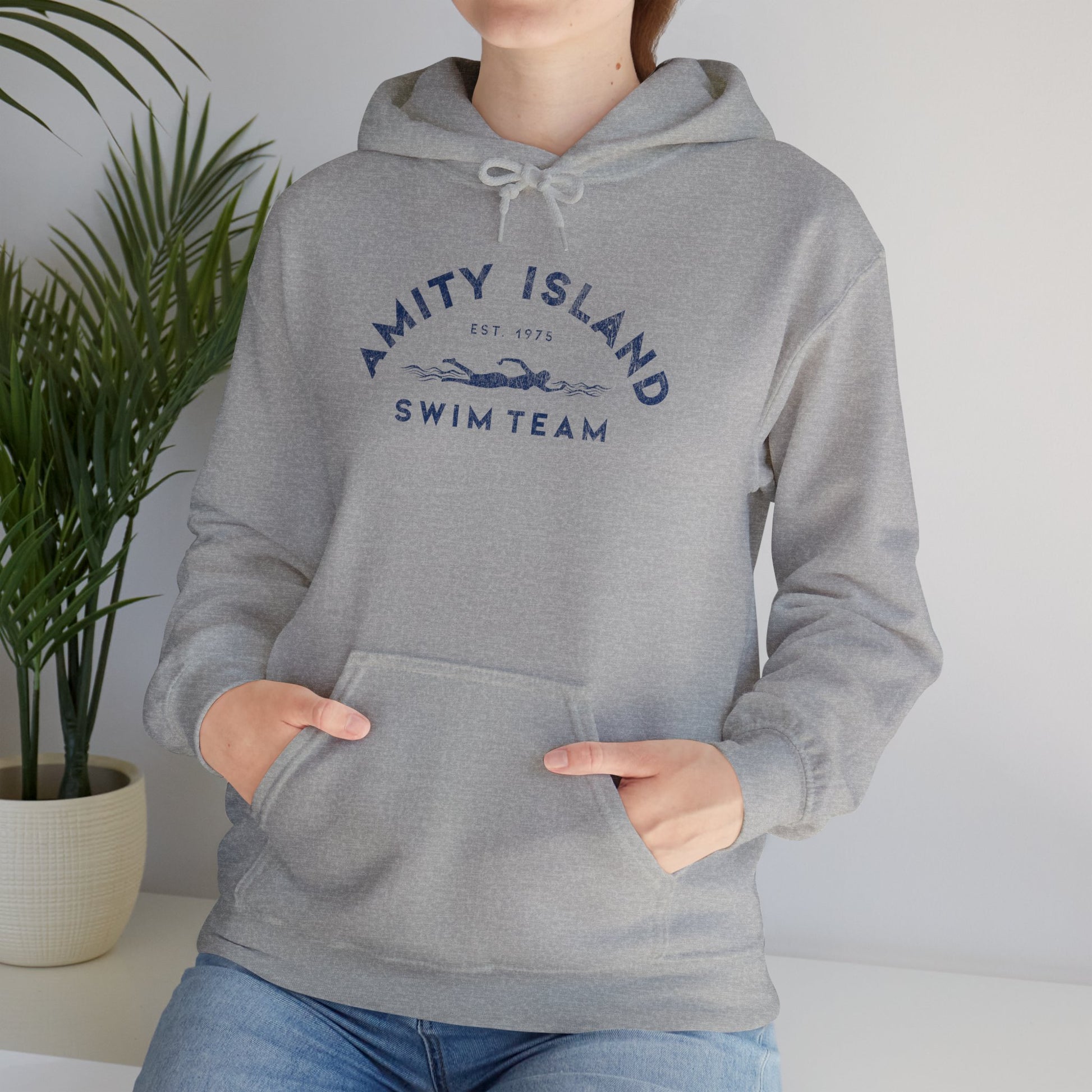 Female model wearing a sport heather grey colored Soft Unisex Hoodie Heavy Blend Hooded Sweatshirt featuring an Amity Island Swim Team logo graphic, designed by ScriptedBrand, and  inspired by the movie Jaws in a vintage distressed style.