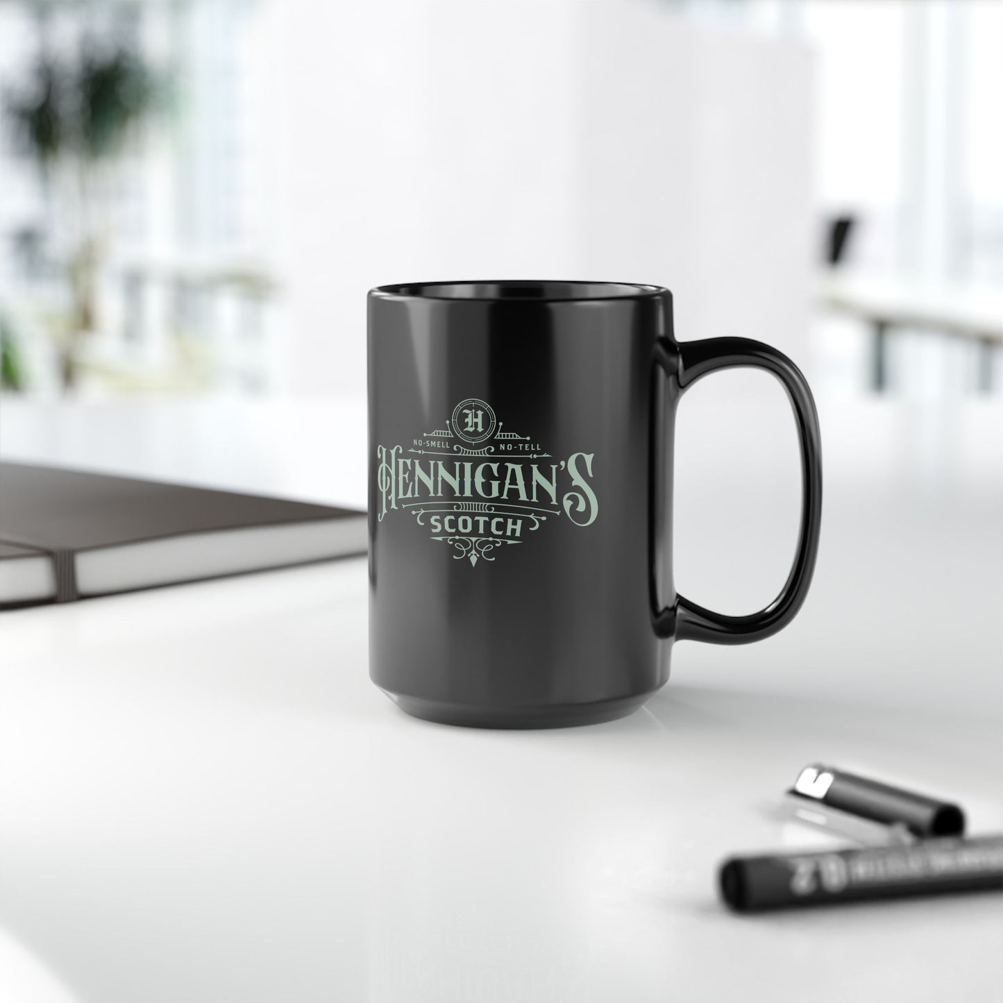 Black mug with 'Hennigan's Scotch' text on a white surface with pens in the background