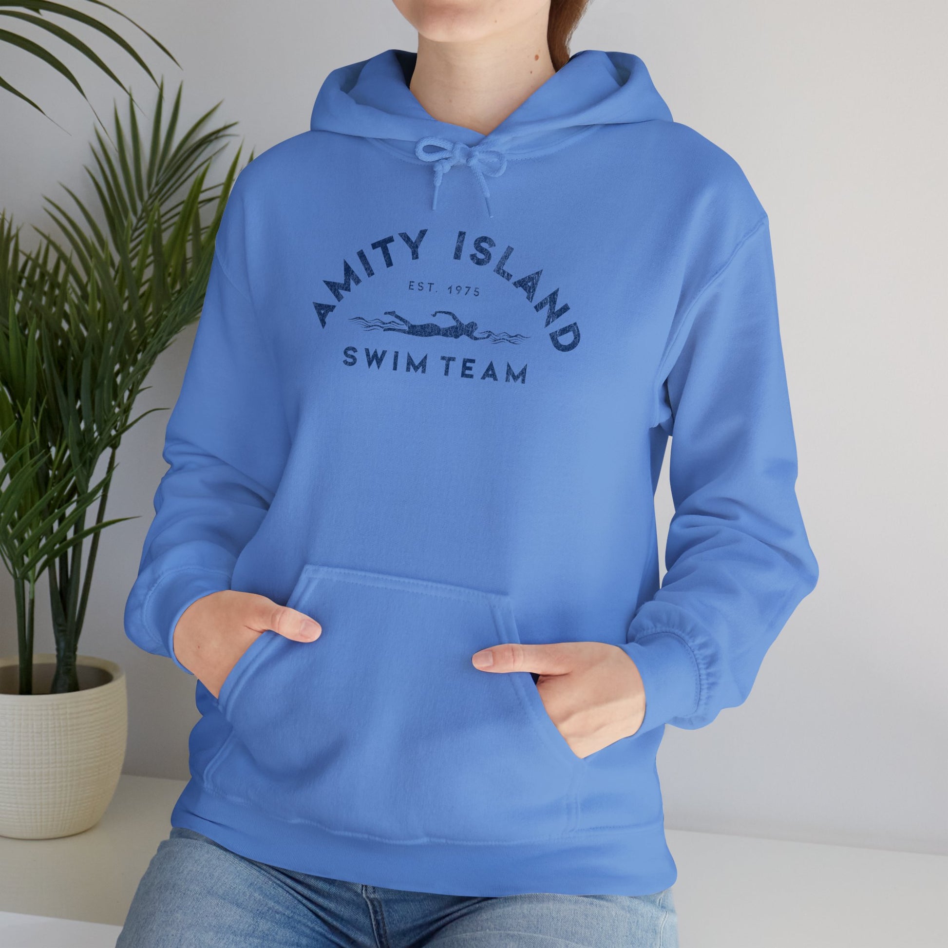 Female model wearing a Carolina blue colored Soft Unisex Hoodie Heavy Blend Hooded Sweatshirt featuring an Amity Island Swim Team logo graphic, designed by ScriptedBrand, and  inspired by the movie Jaws in a vintage distressed style.