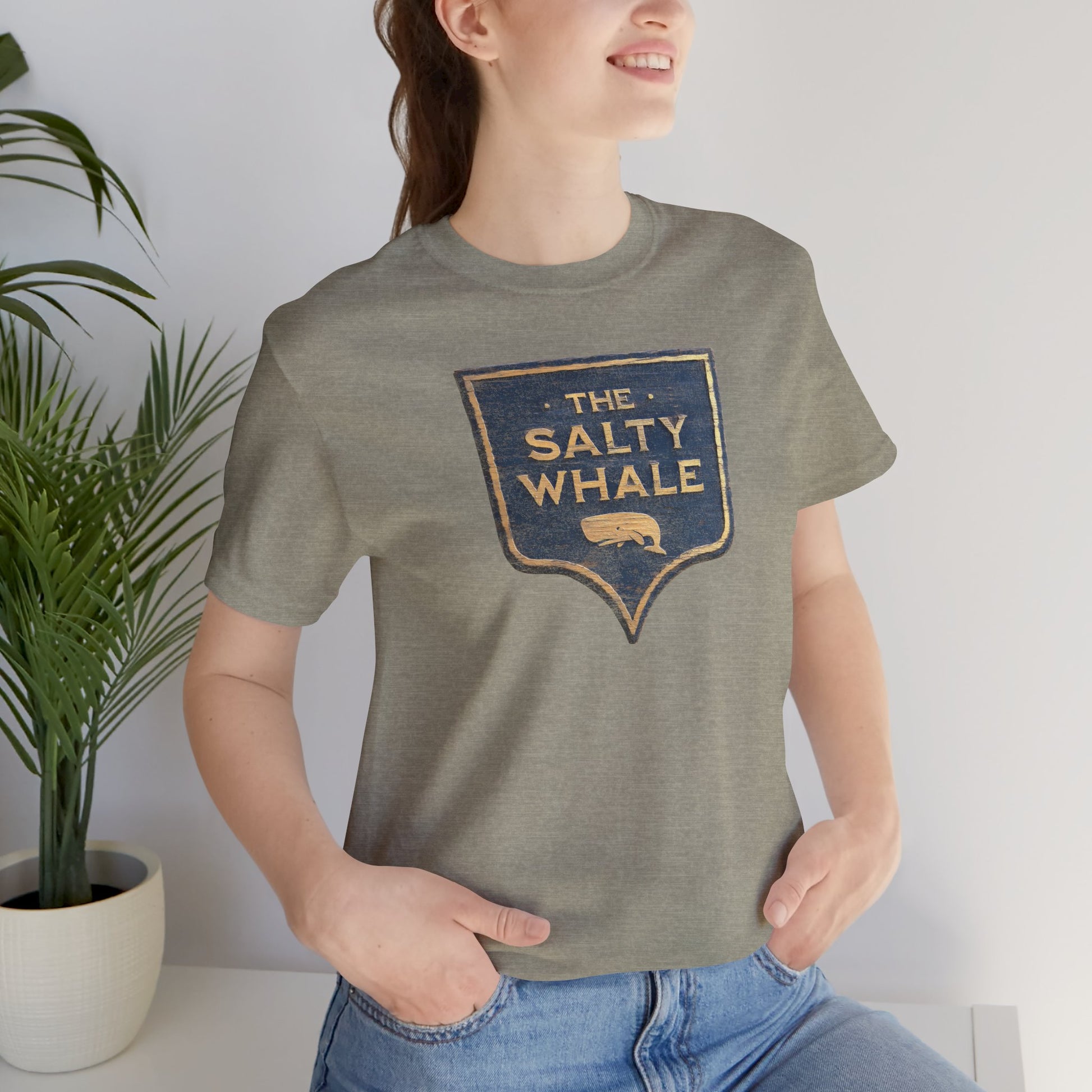 Woman wearing Heather stone colored 'The Salty Whale' weathered carved-wood pub sign t-shirt inspired by Widow's Bay Apple TV series, heather Bella+Canvas tee