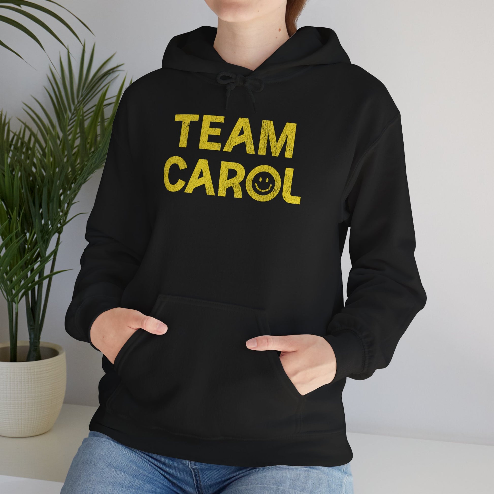 Woman wearing black unisex Heavy Blend pullover hoodie sweatshirt with TEAM CAROL vintage distressed smiley graphic