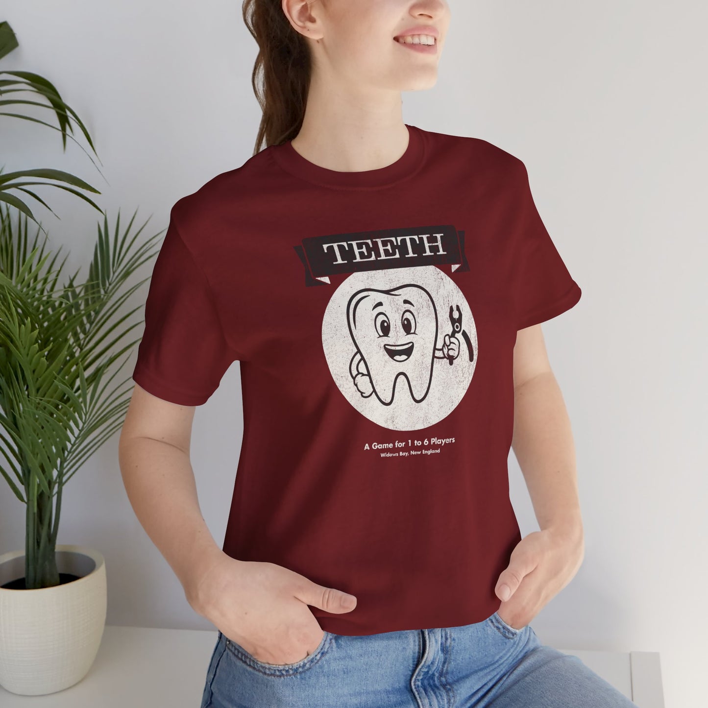 Woman wearing Red Bella+Canvas tee with the vintage TEETH board game box cover from Widow's Bay, a smiling tooth holding pliers