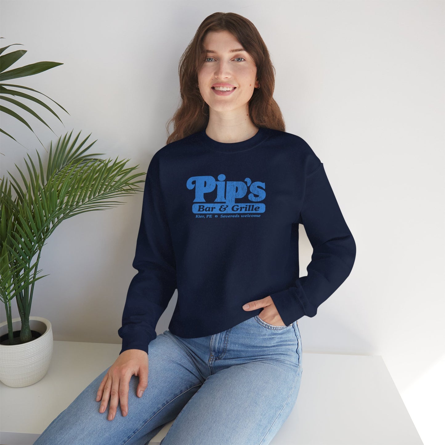 Female model wearing a navy blue soft Gildan 18000 sweatshirt featuring the Pips Bar and Grill logo Severeds Welcome from the TV show Severance in a vintage distressed ink-stamped worn-in style.
