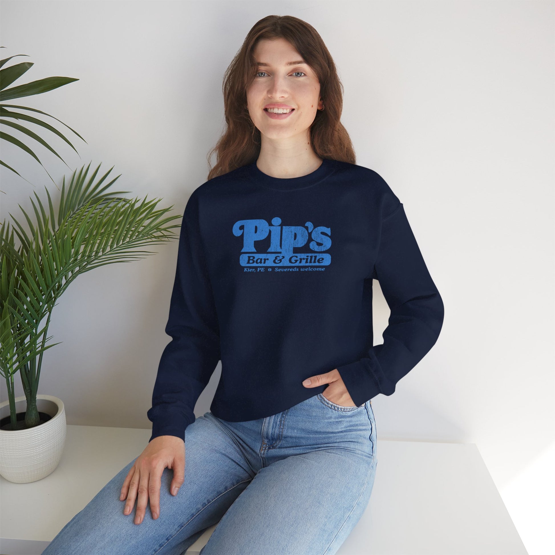 Female model wearing a navy blue soft Gildan 18000 sweatshirt featuring the Pips Bar and Grill logo Severeds Welcome from the TV show Severance in a vintage distressed ink-stamped worn-in style.